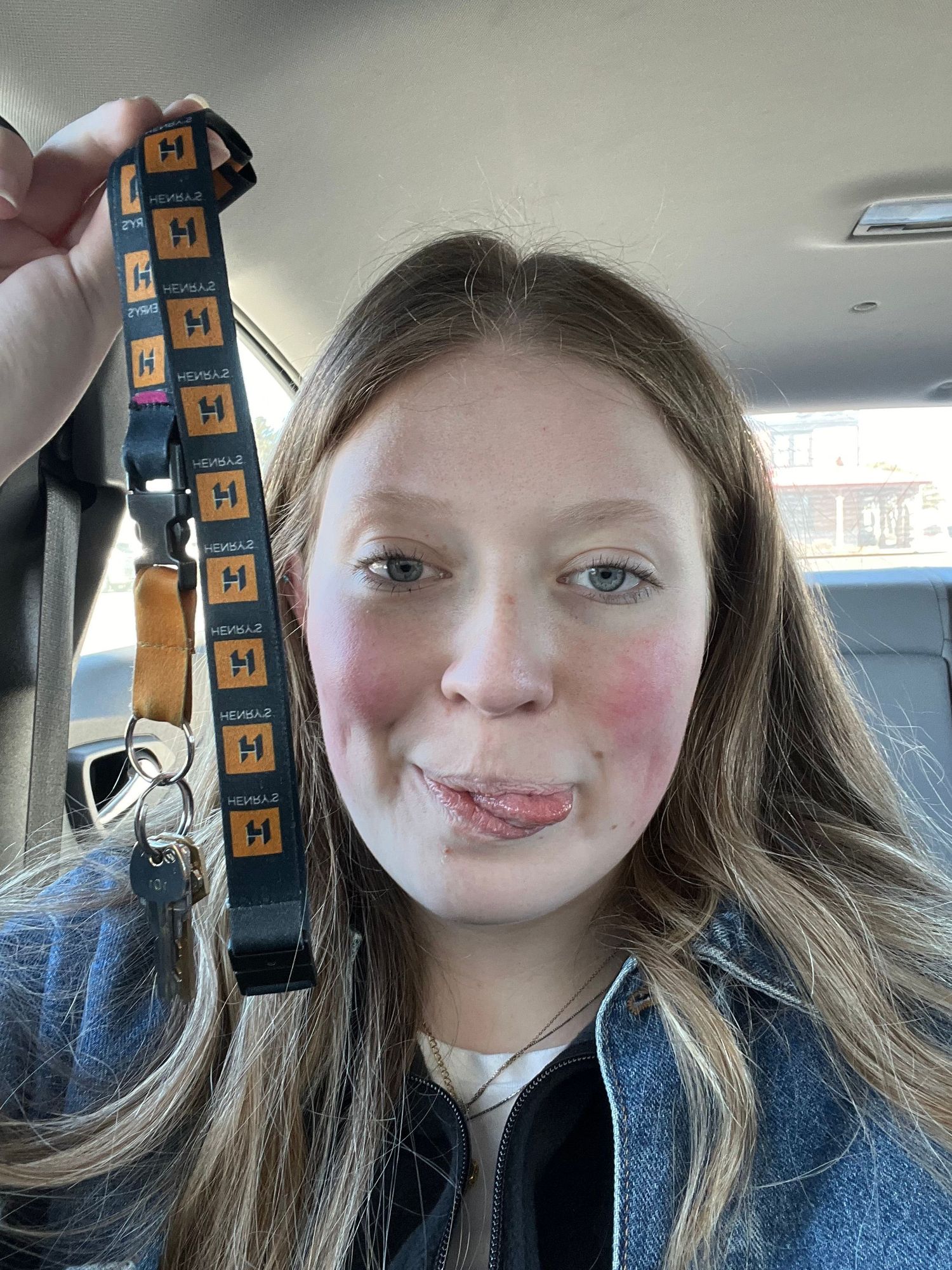 A person in a blue denim jacket holds up a dangling orange and black lanyard while taking selfies in a car.