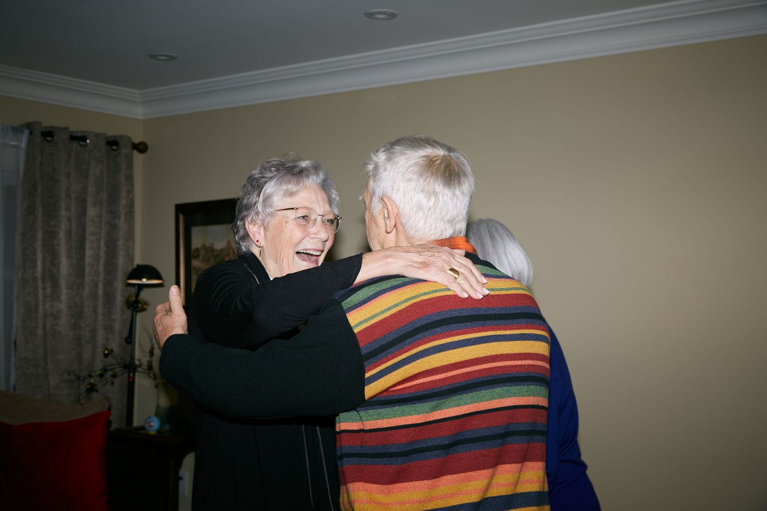 Three elderly people dance together in a living room with curtains and beige walls in the background.