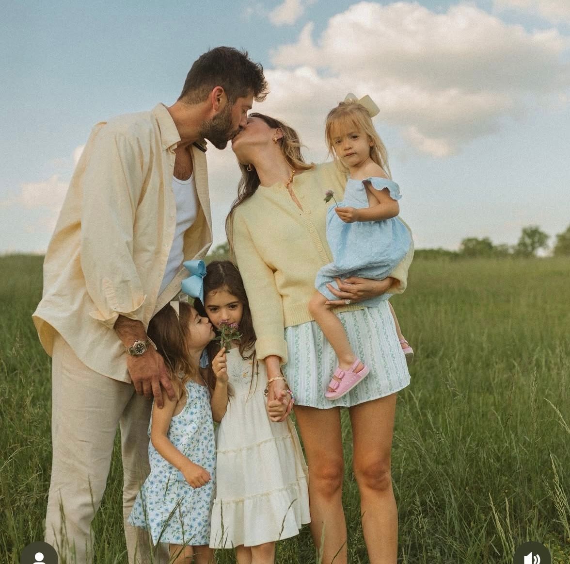A family of four poses in a green field, parents kissing while holding two young daughters dressed in pastel outfits.