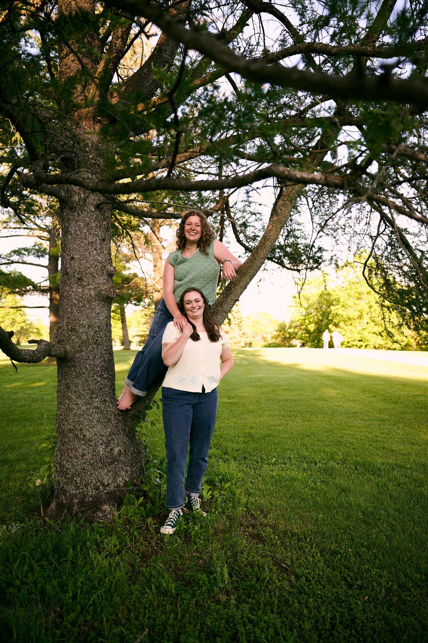 Two young women smiling outdoors near a large tree in a green park, one sitting in the branches, one standing below.