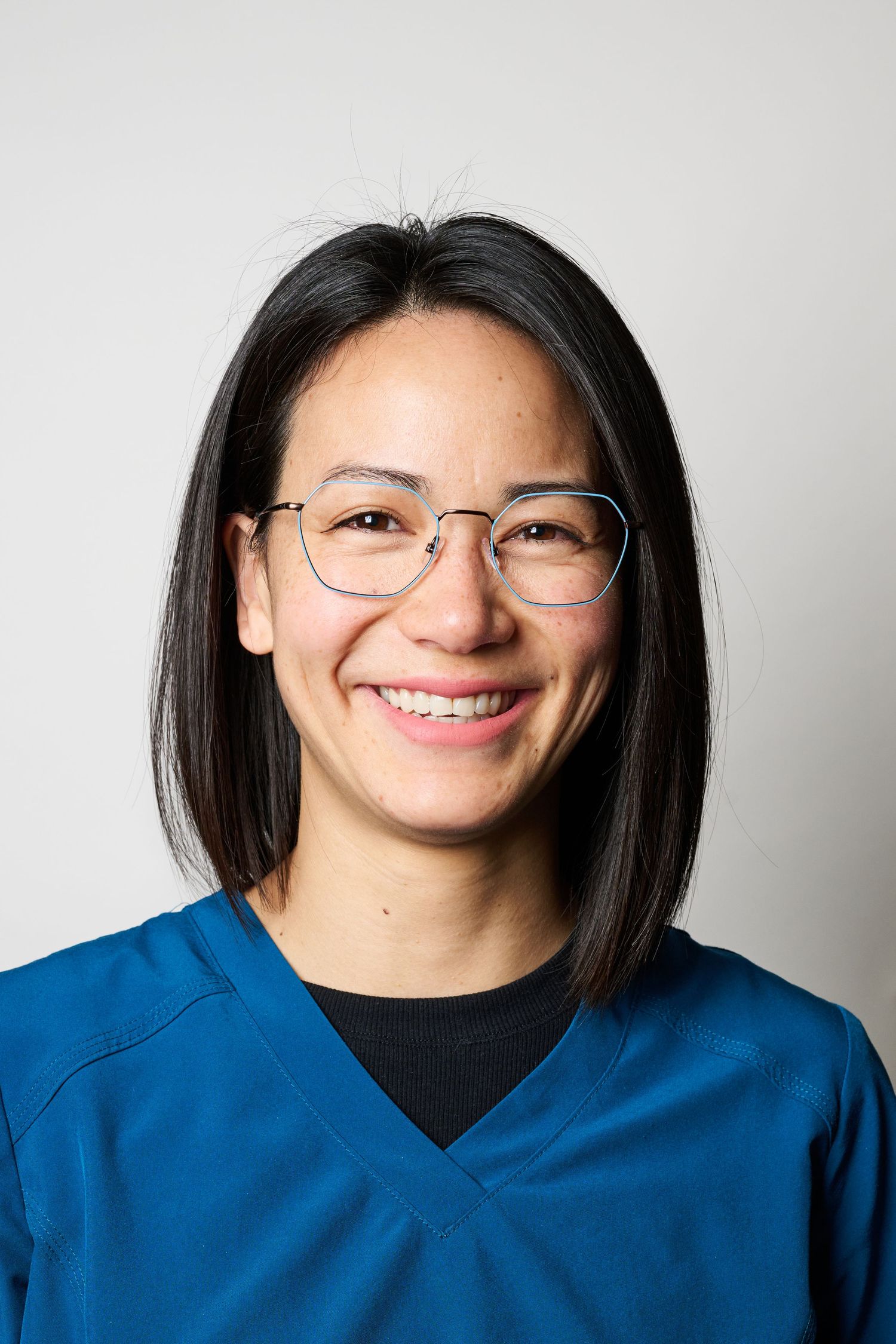 A person in a blue scrub top smiles warmly while wearing glasses against a light background.