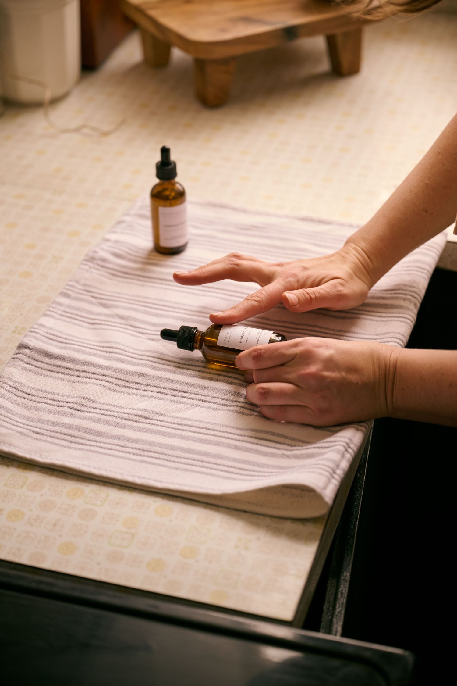 Hands handling dropper bottles on a white striped cloth with essential oils and wooden decor visible.