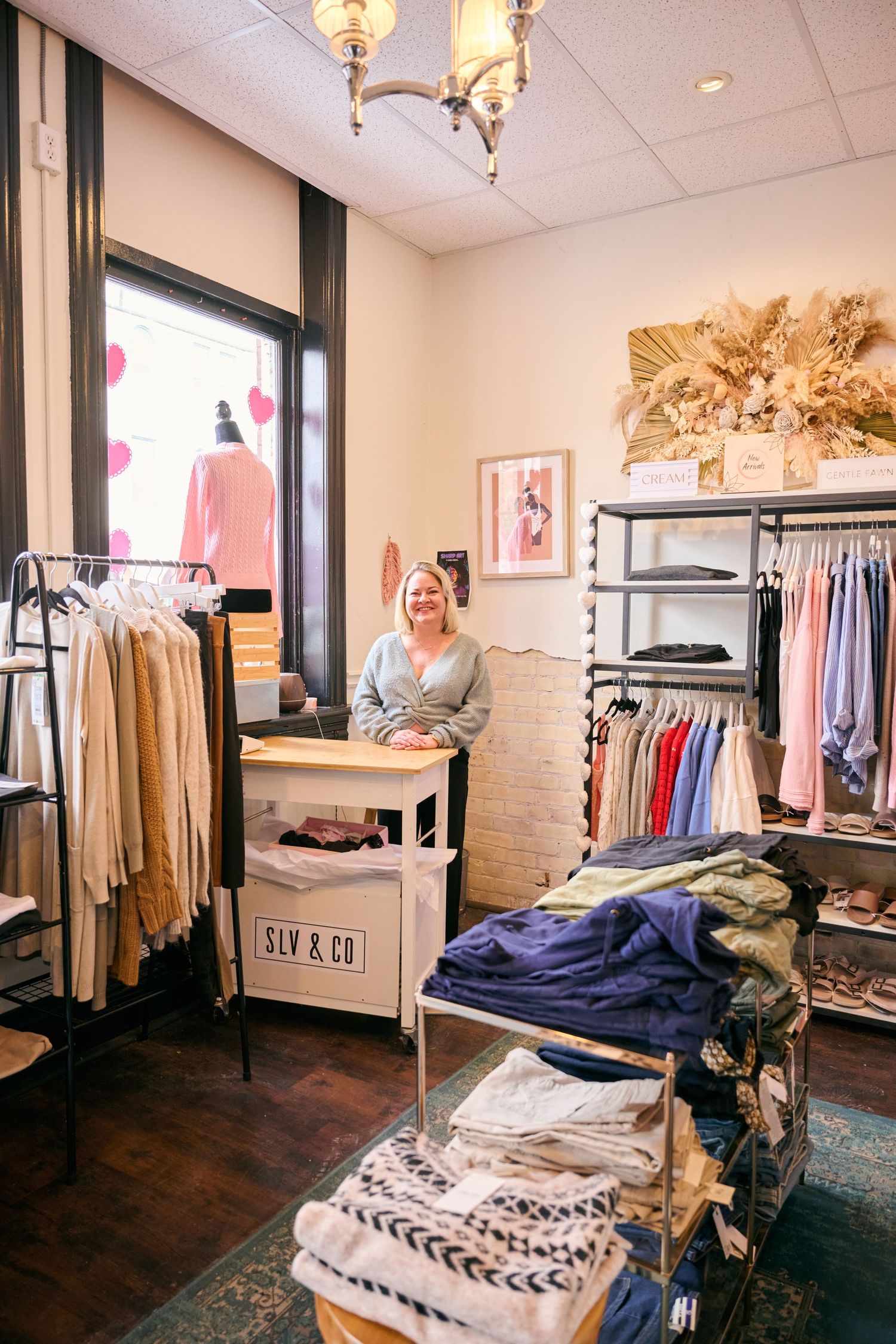 Cozy boutique clothing store interior with racks of apparel, a sales counter, and warm lighting from a chandelier.