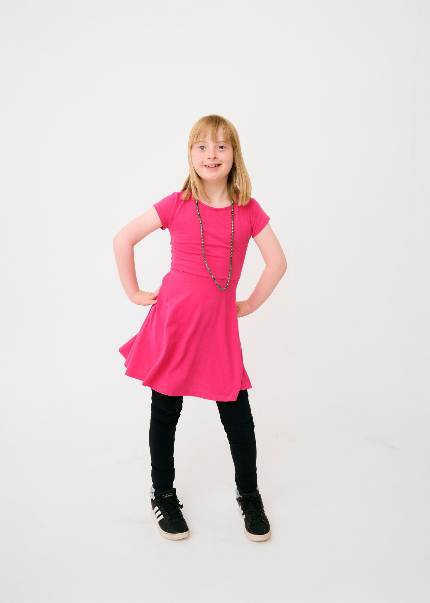 A young dancer in a bright pink dress and black leggings strikes a confident pose against a white background.