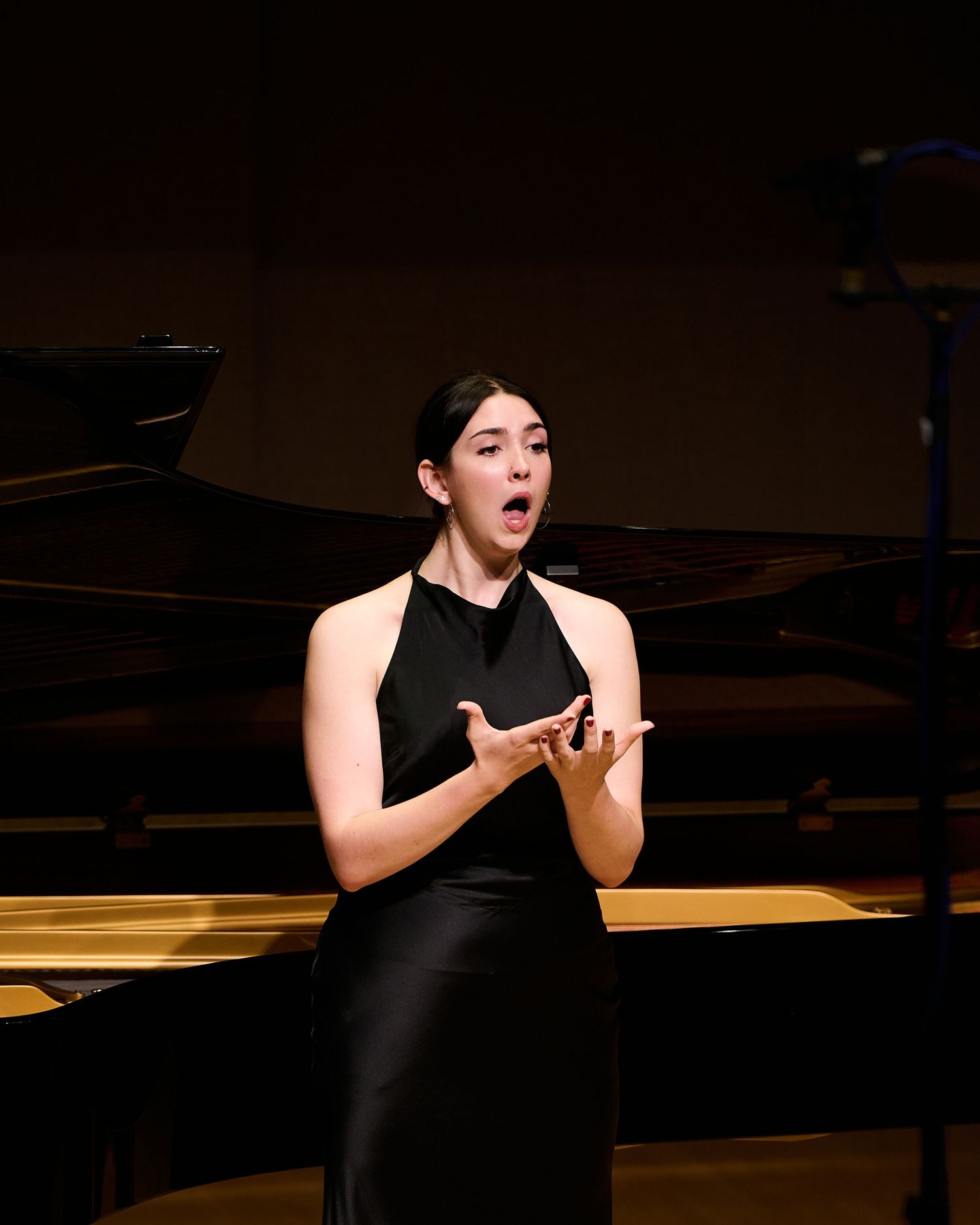 Woman in black dress performing opera on stage near a grand piano.