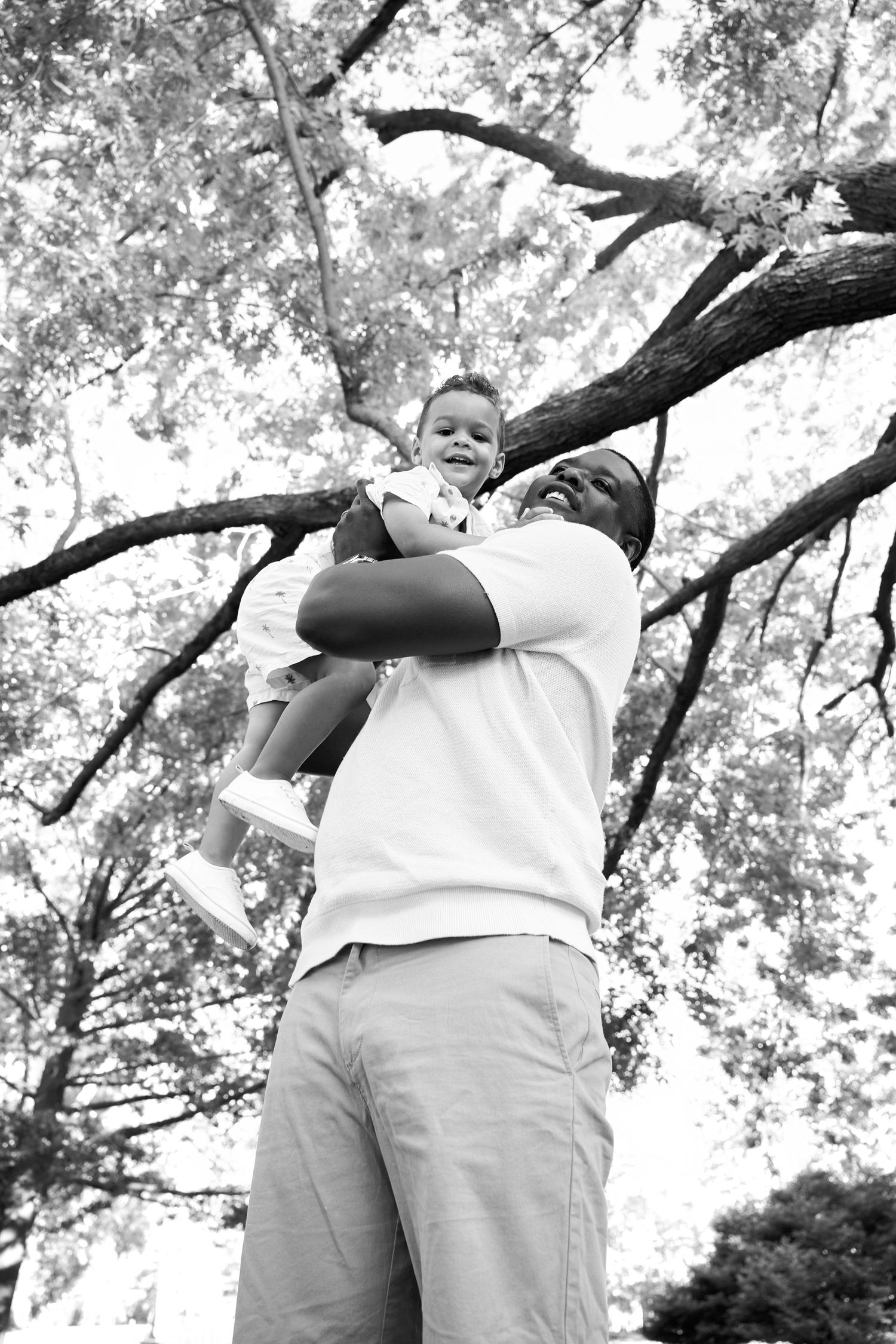 Black and white photo of a father lifting his smiling child outdoors under tree branches in a park.