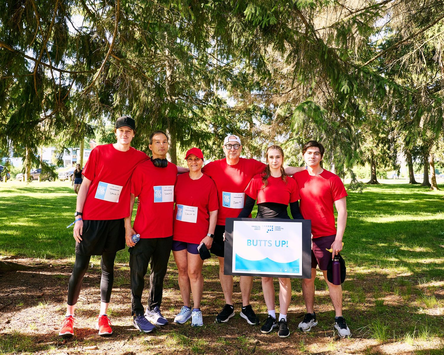 Group of six runners in red shirts posing with a Putts Up sign in a sunny park setting.