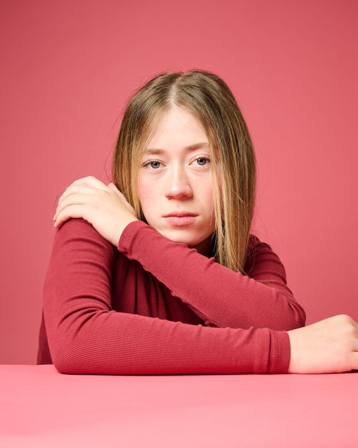 Person wearing burgundy sweater rests head on crossed arms against pink background in contemplative series.