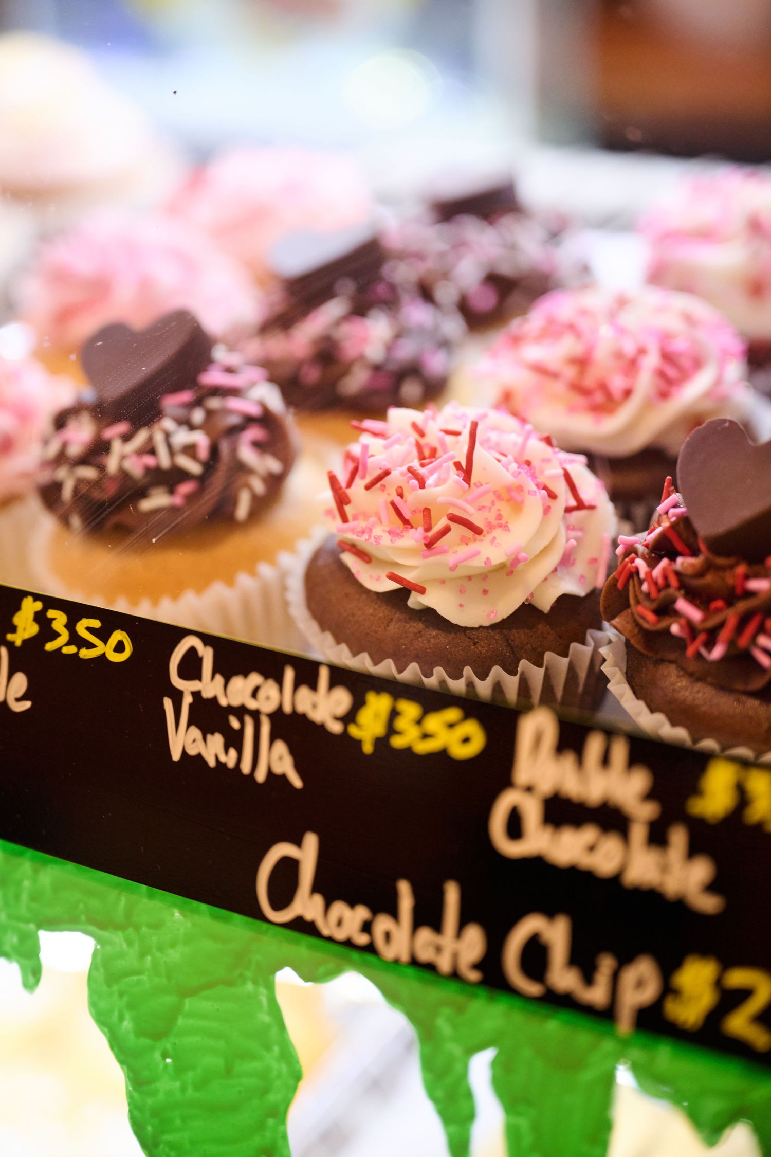 Chocolate and vanilla cupcakes with pink frosting and sprinkles displayed with price tags in a bakery case.