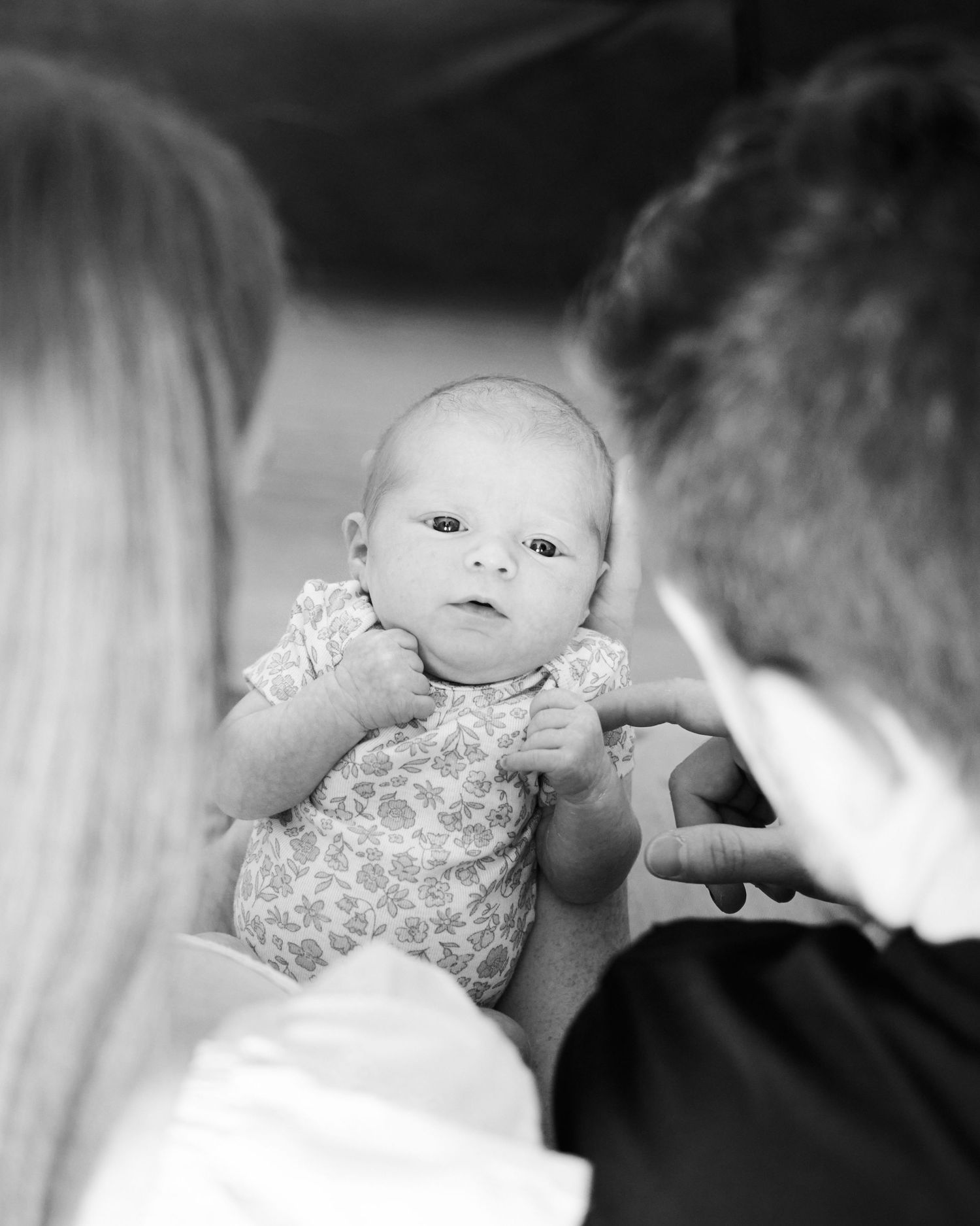 Black and white photo of a newborn baby being held by two parents, gazing curiously at the camera.