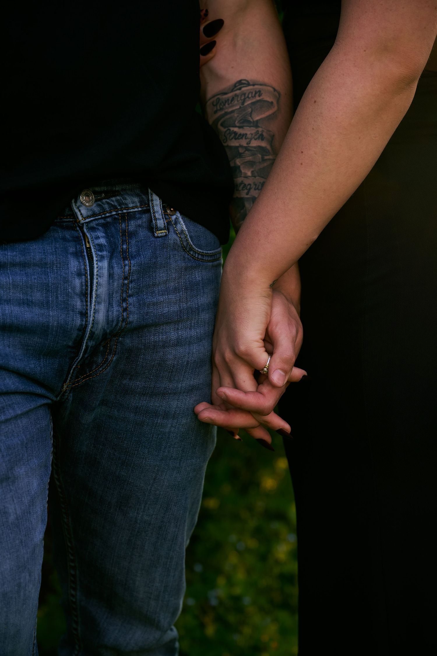 Two people holding hands, one with a tattoo on their arm, wearing blue jeans outdoors.