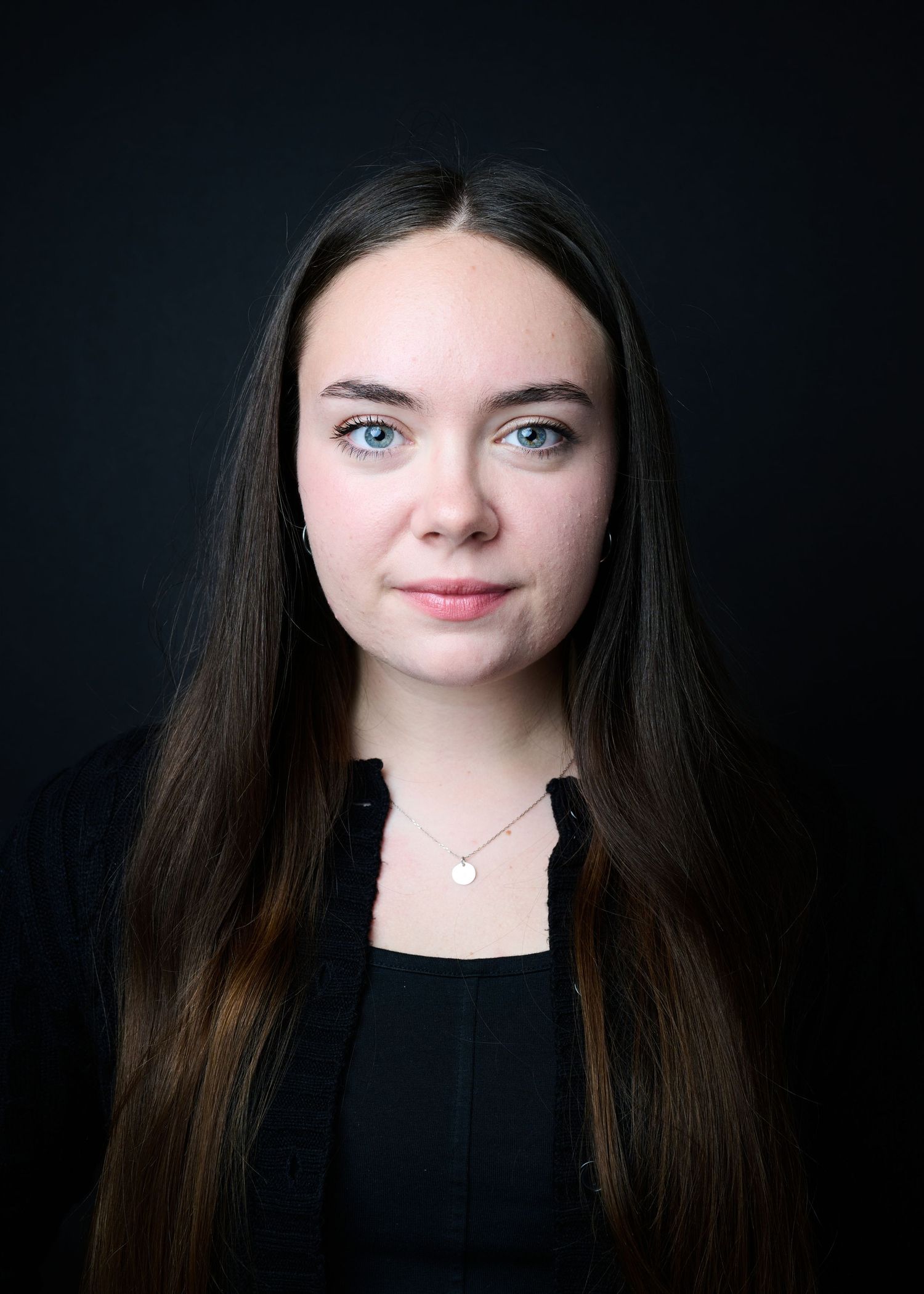 Portrait against dark background showing long dark hair and black top with delicate heart necklace.