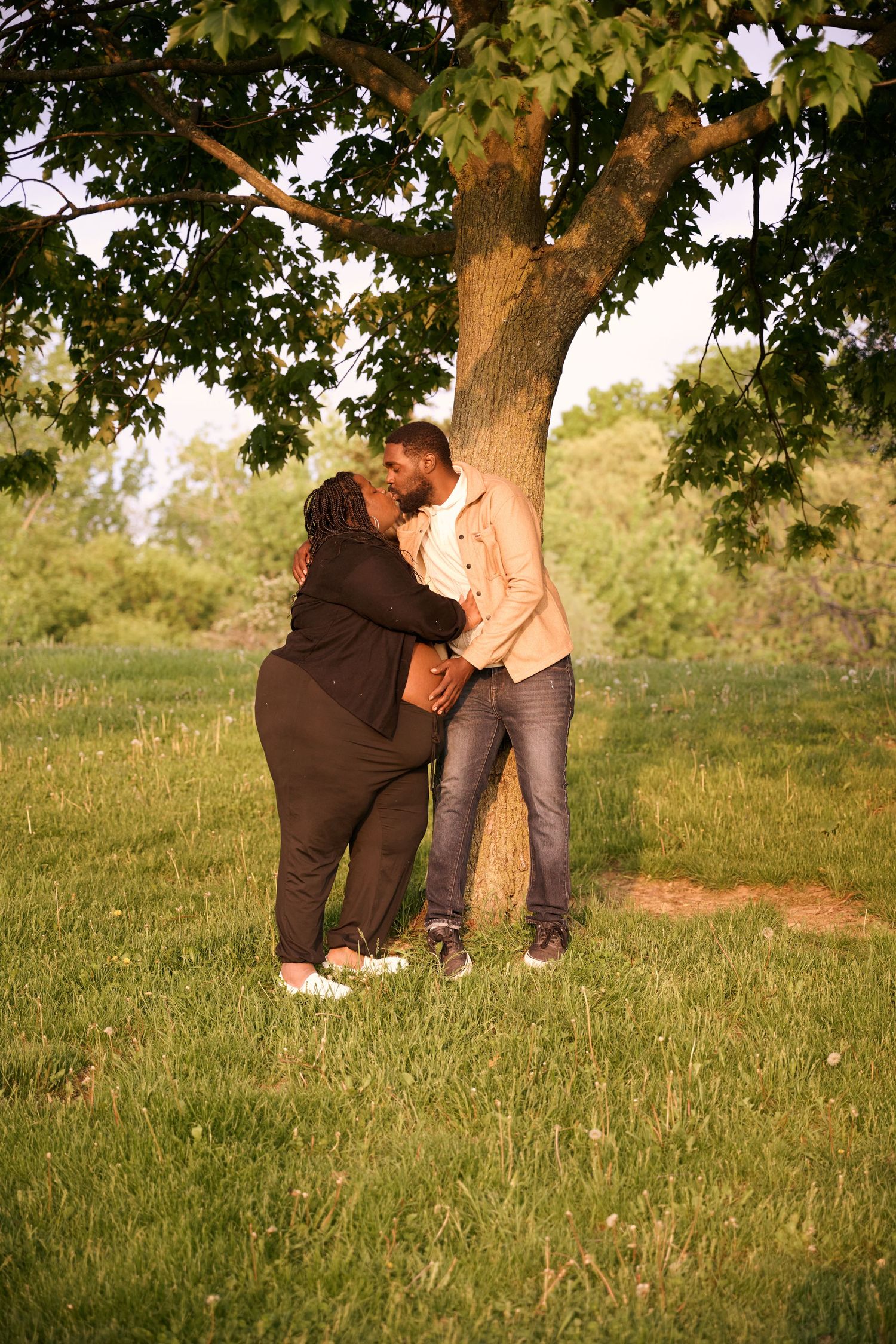 A couple shares an intimate moment beneath a large tree in a sunlit grassy field during golden hour.