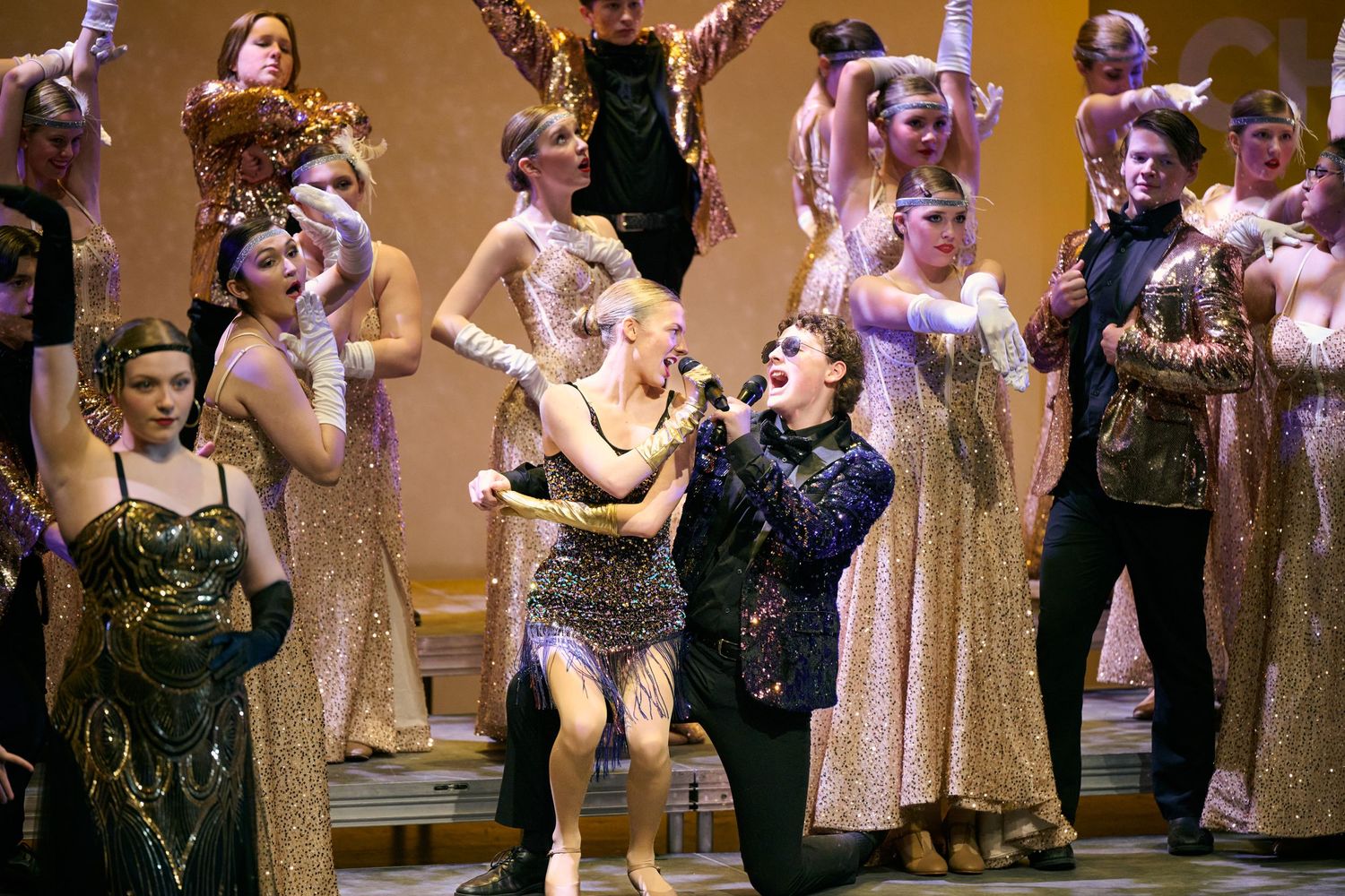 Performers in glittering vintage costumes dance and pose during a theatrical stage production with golden lighting.