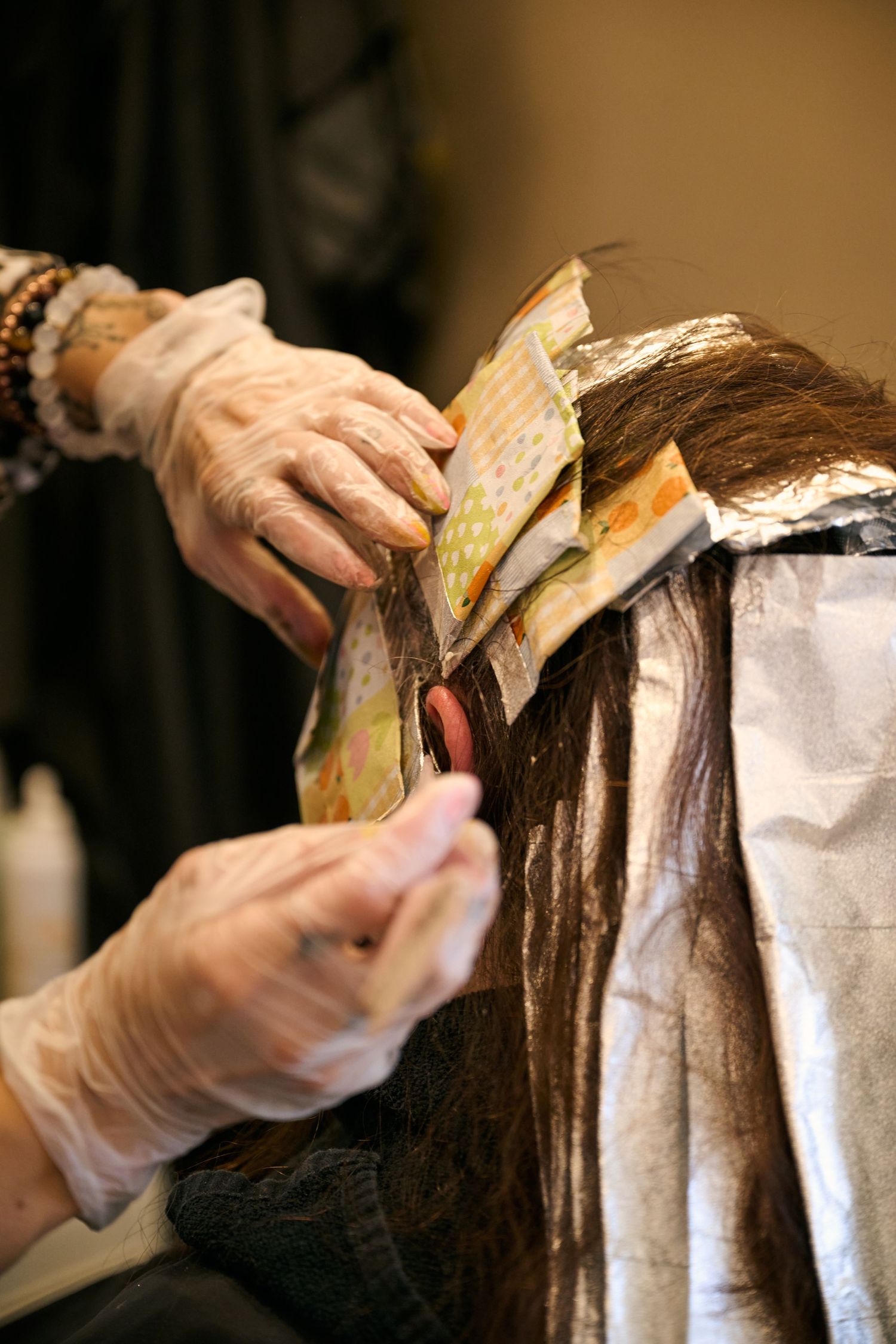 A hairdresser's gloved hands applying foil highlights to long dark hair during a salon coloring treatment.