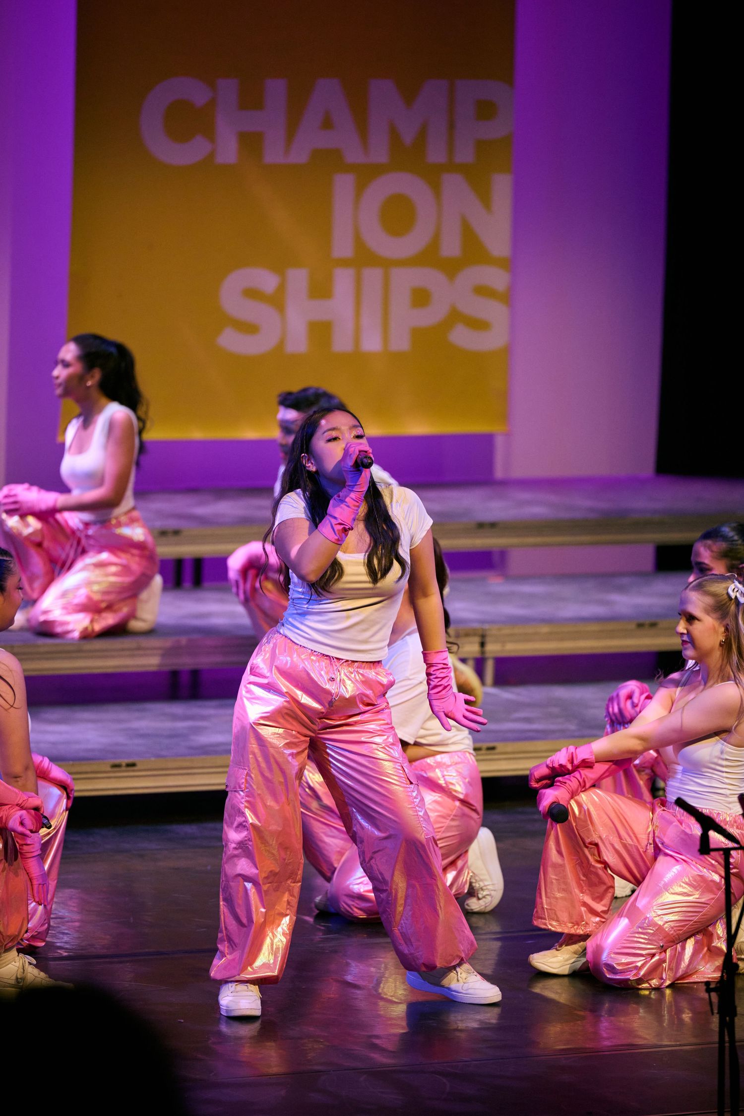 Dancers in pink and white outfits perform energetic hip-hop choreography on a purple-lit stage.