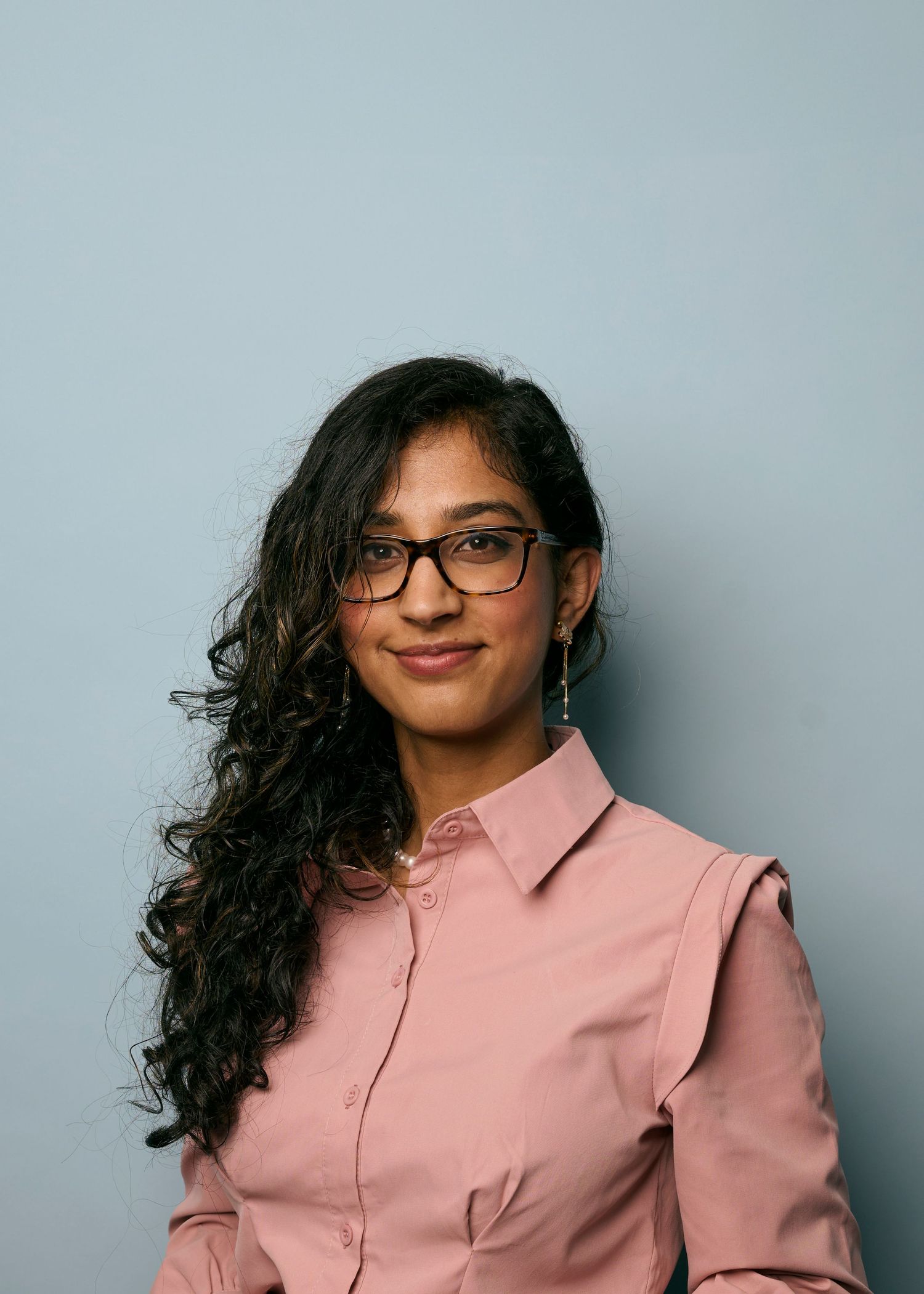 A person with long dark curly hair wearing glasses and a pink button-up shirt poses in a series of photos.