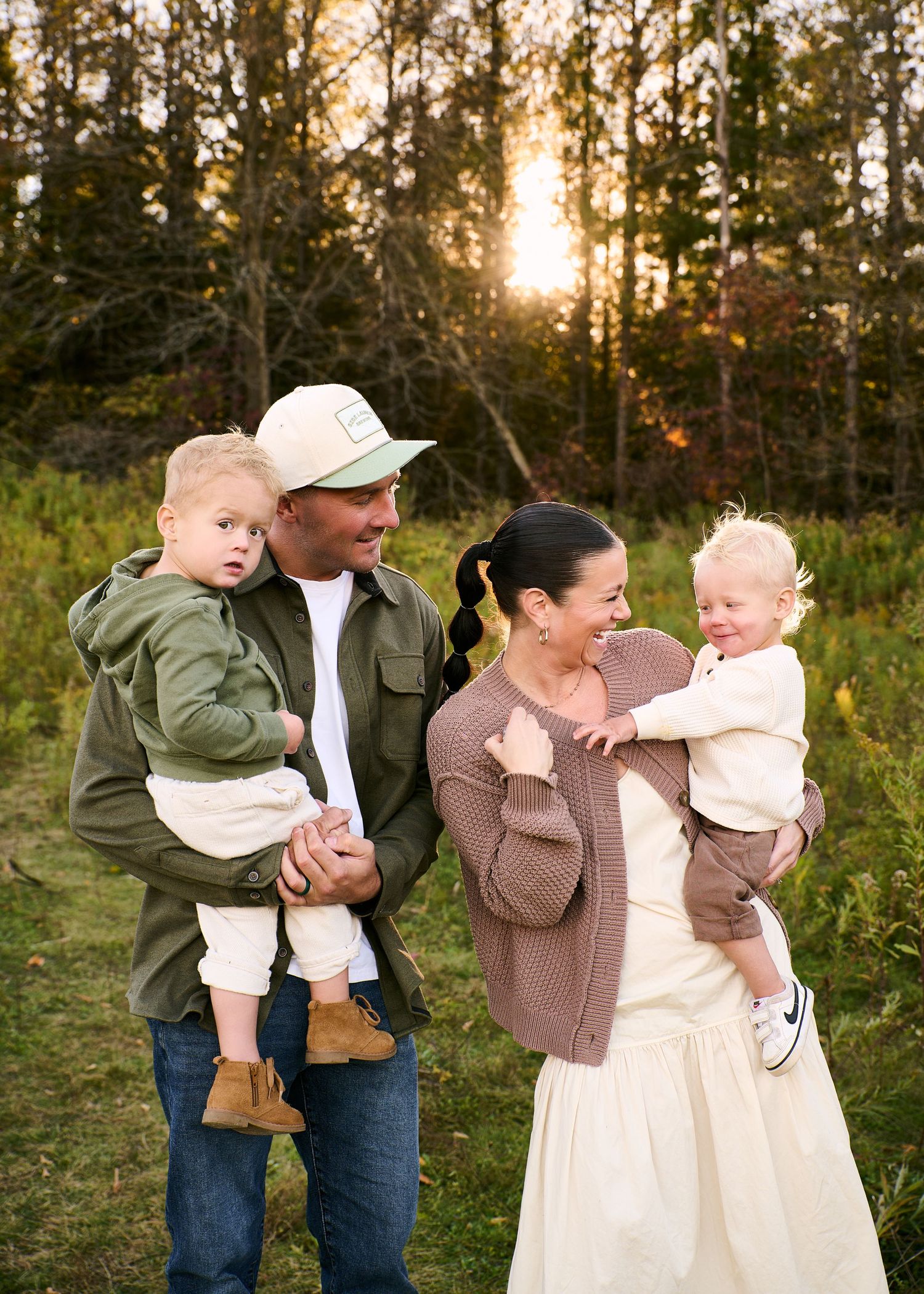 Smiling family of four holding two toddlers outdoors in a grassy field at golden hour, with sunlight glowing through tall trees behind them.