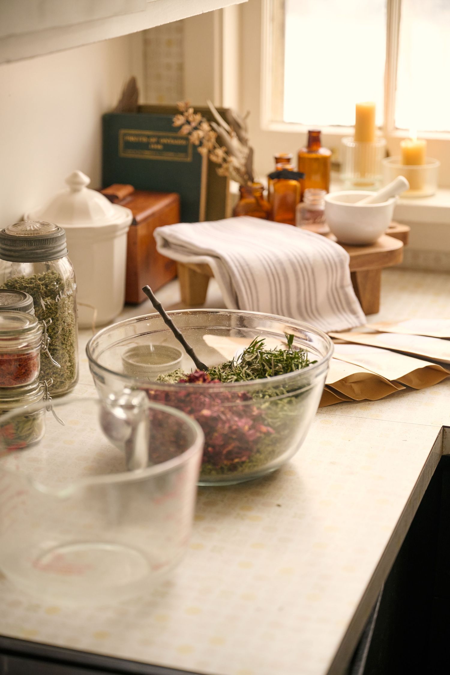 A rustic kitchen counter with glass bowls, mason jars of herbs, and white linens in warm natural lighting.