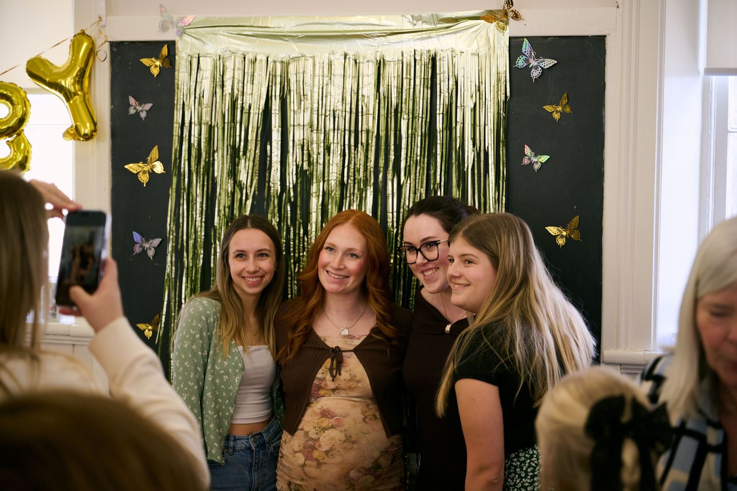 A group poses together at a party with a green tinsel backdrop and yellow butterfly decorations on the wall.