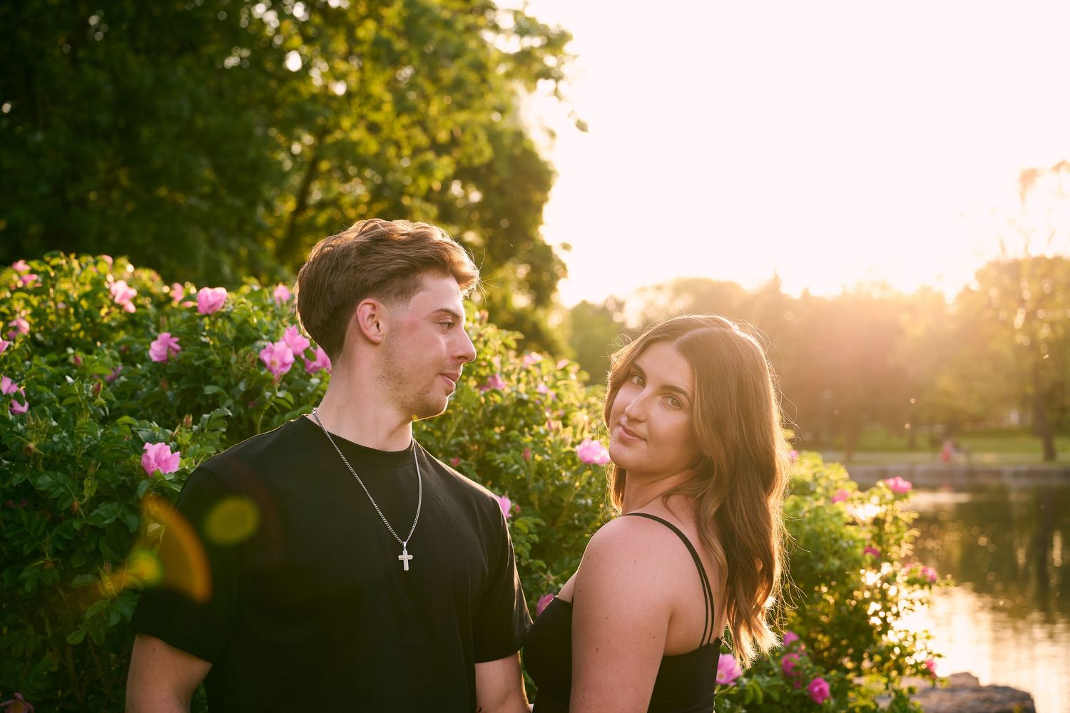 Couple posing together at golden hour near a pond surrounded by blooming pink roses and lush green trees.
