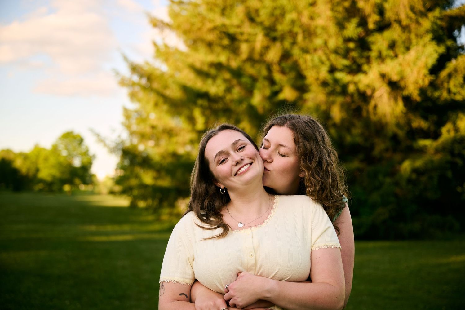 Two young women share a tender moment outdoors, one kissing the other's cheek while embracing in a sunny park.