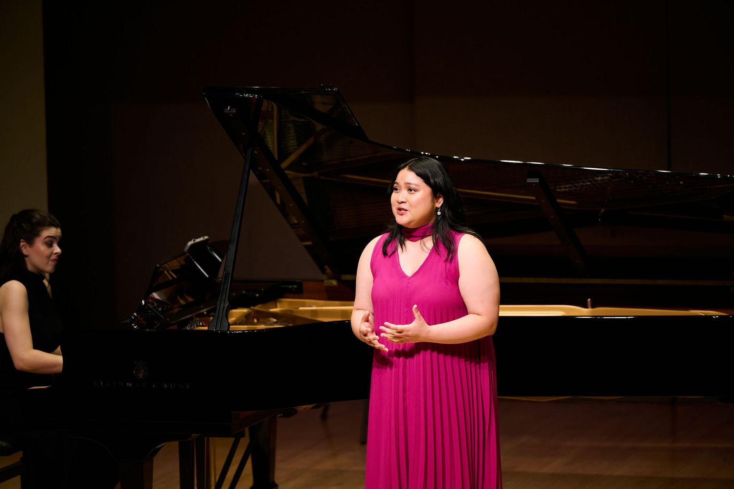 Woman in pink dress singing on stage with grand piano accompanist in background at formal recital.