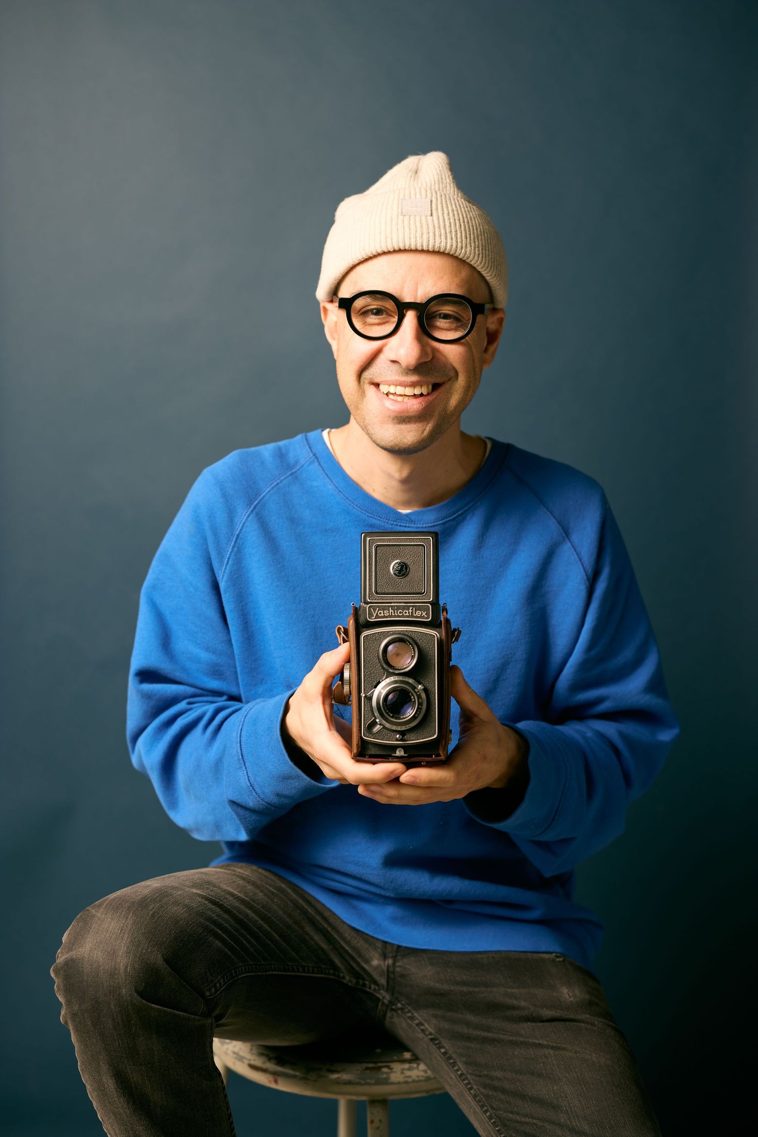 Photographer in blue sweater holding vintage twin lens reflex camera against dark studio backdrop in multiple poses.