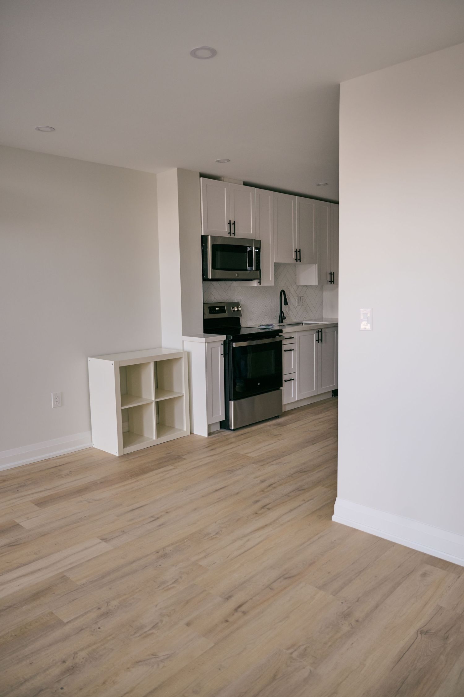 Modern apartment interior with light hardwood floors, white cabinets, and built-in appliances in an open concept kitchen layout.