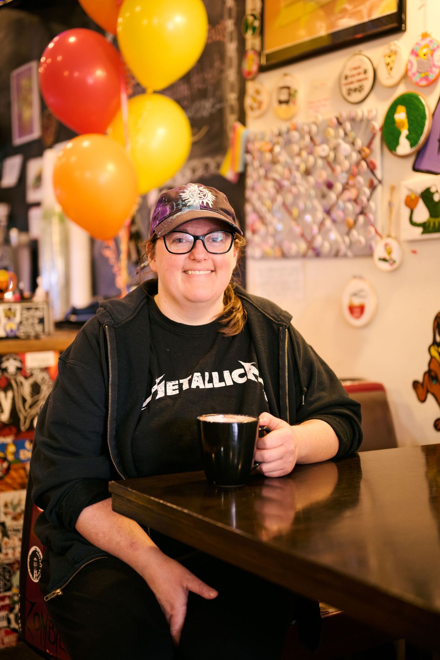 Someone relaxes at a pub with colorful balloons wearing a Metallica shirt and baseball cap while enjoying a dark beer.
