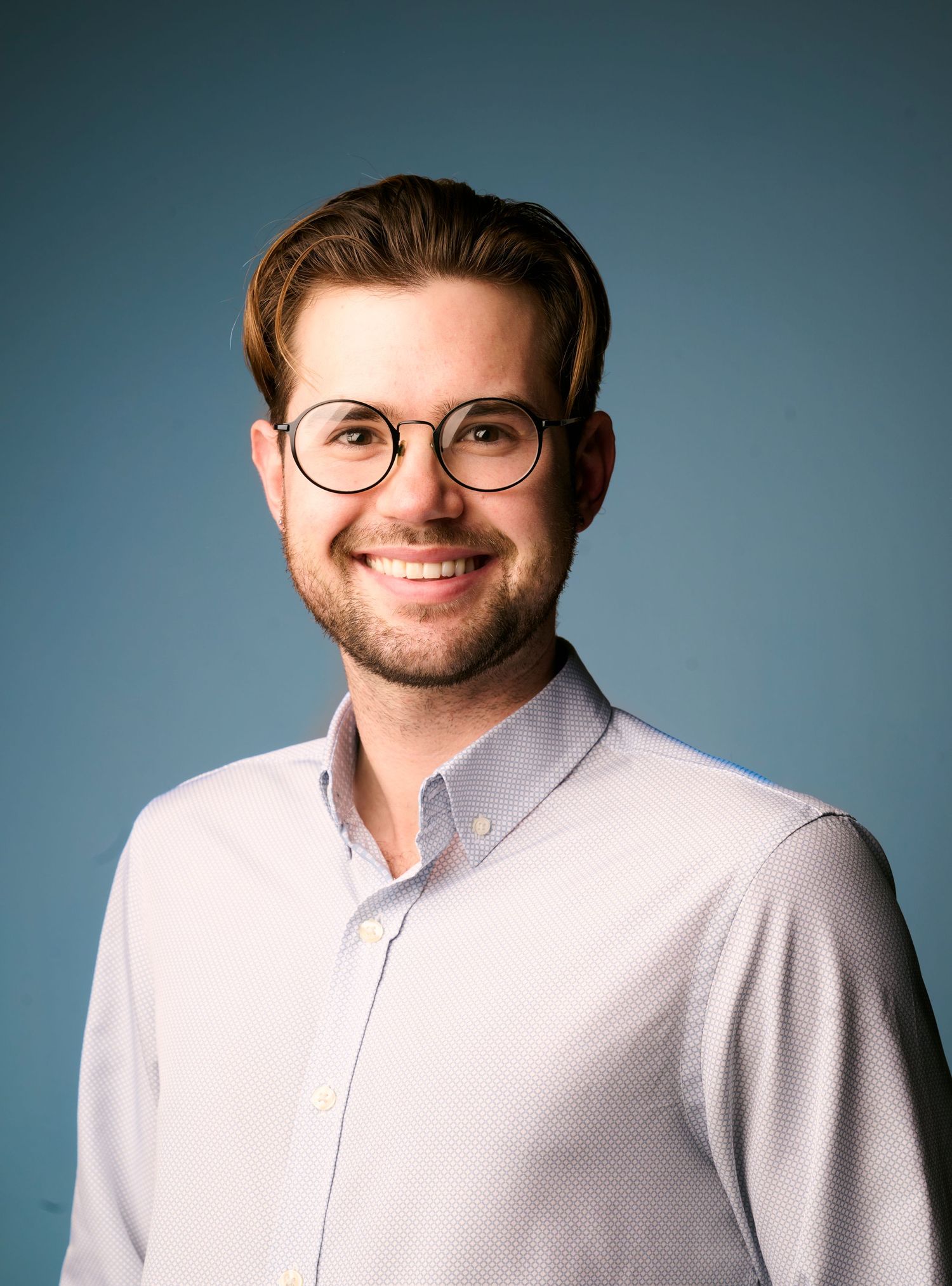 Professional portrait of a person in a light button-down shirt and round glasses against a blue backdrop.