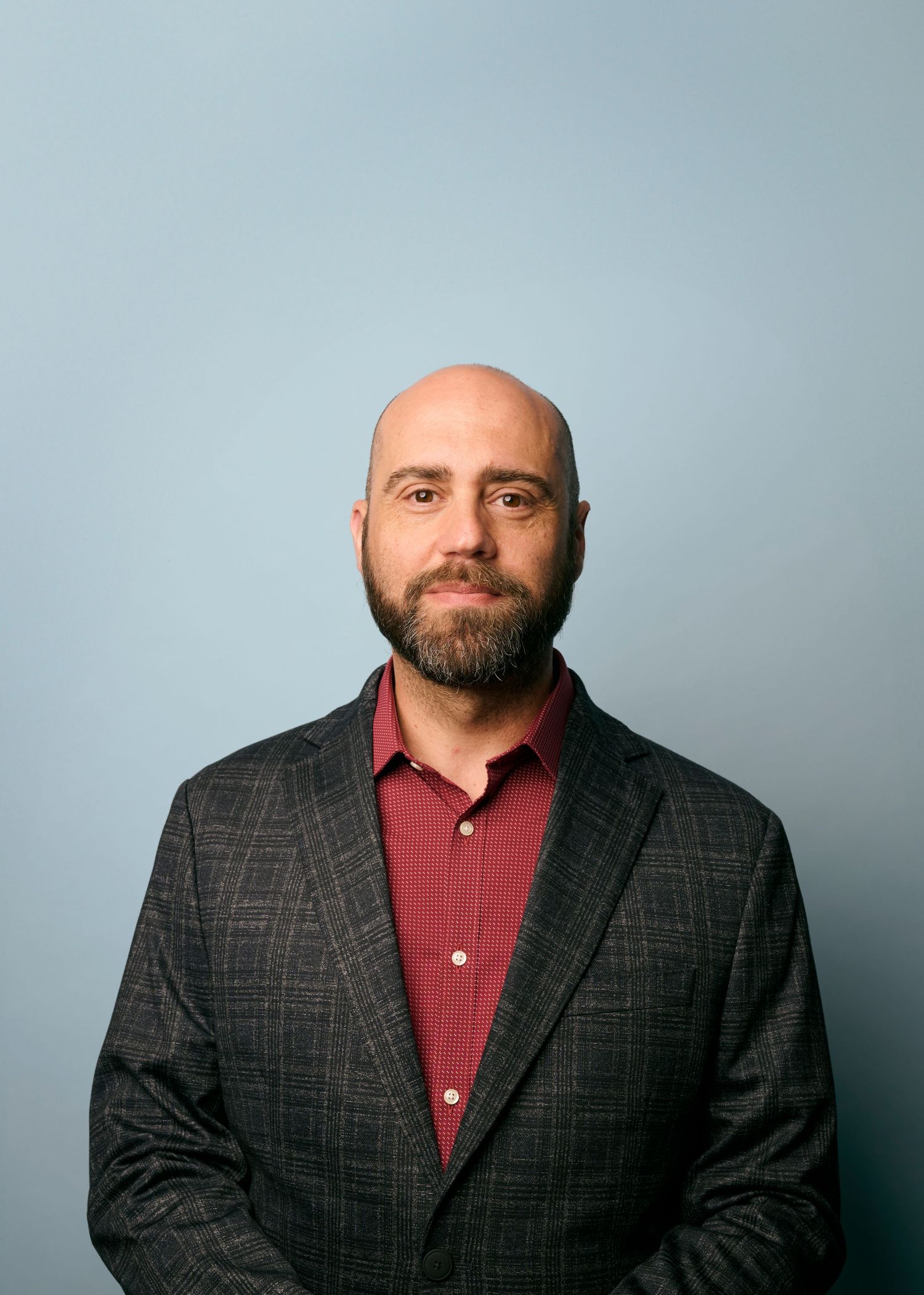 Professional headshot of a bald person wearing a dark plaid blazer and burgundy shirt against a light background.
