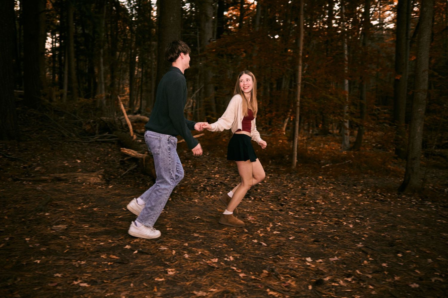 Two people dance playfully in a forest setting during golden hour as autumn leaves cover the ground.