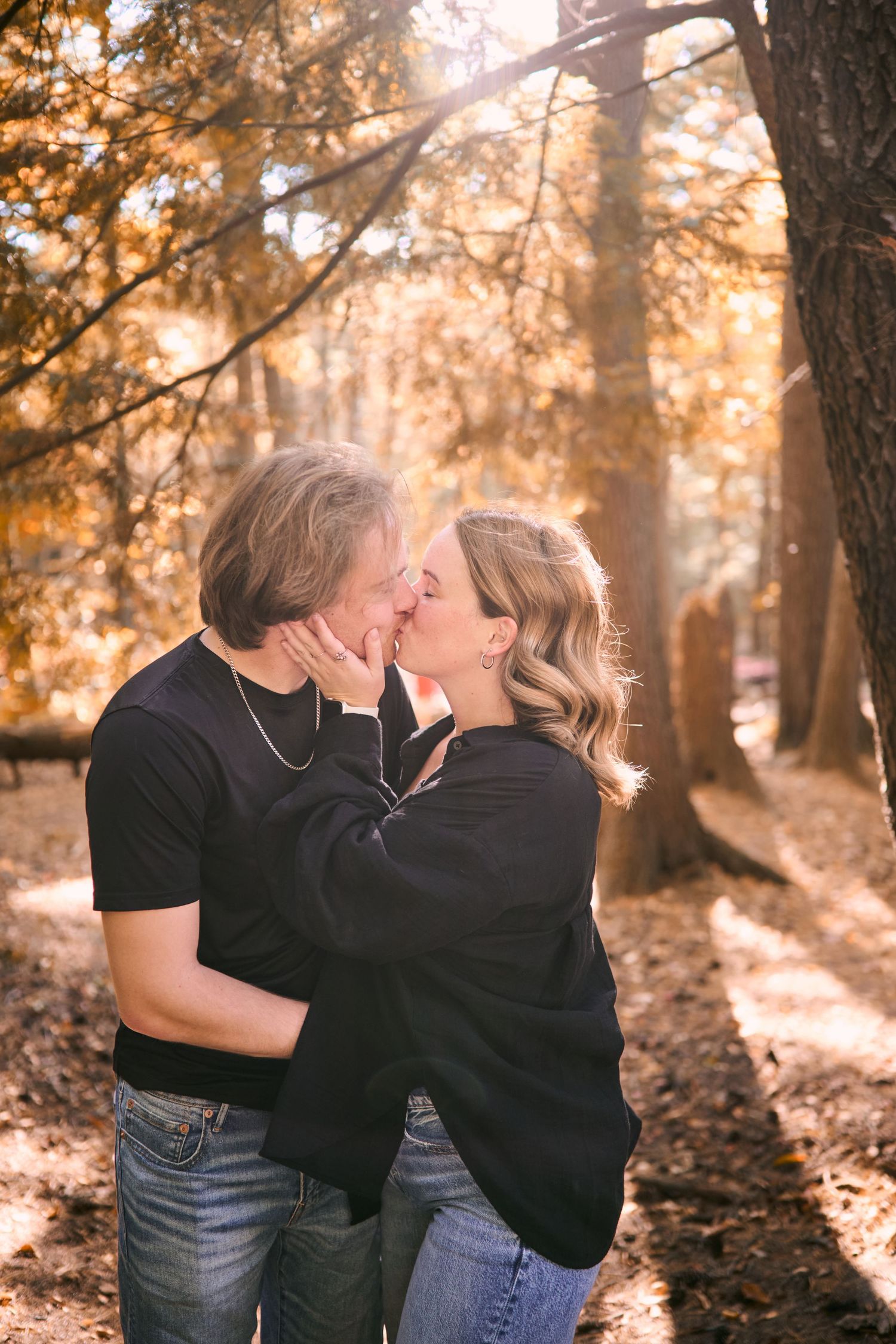 A romantic couple shares a tender kiss in a sunlit autumn forest setting with golden light filtering through the trees.
