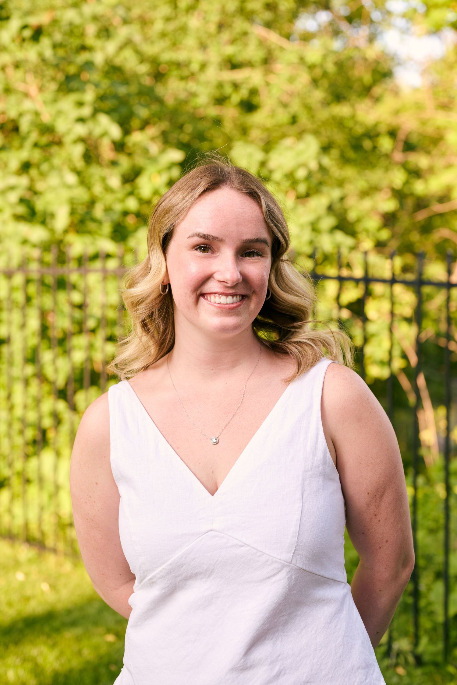A summer portrait of a person in a white sleeveless dress standing outdoors with lush green foliage in the background.