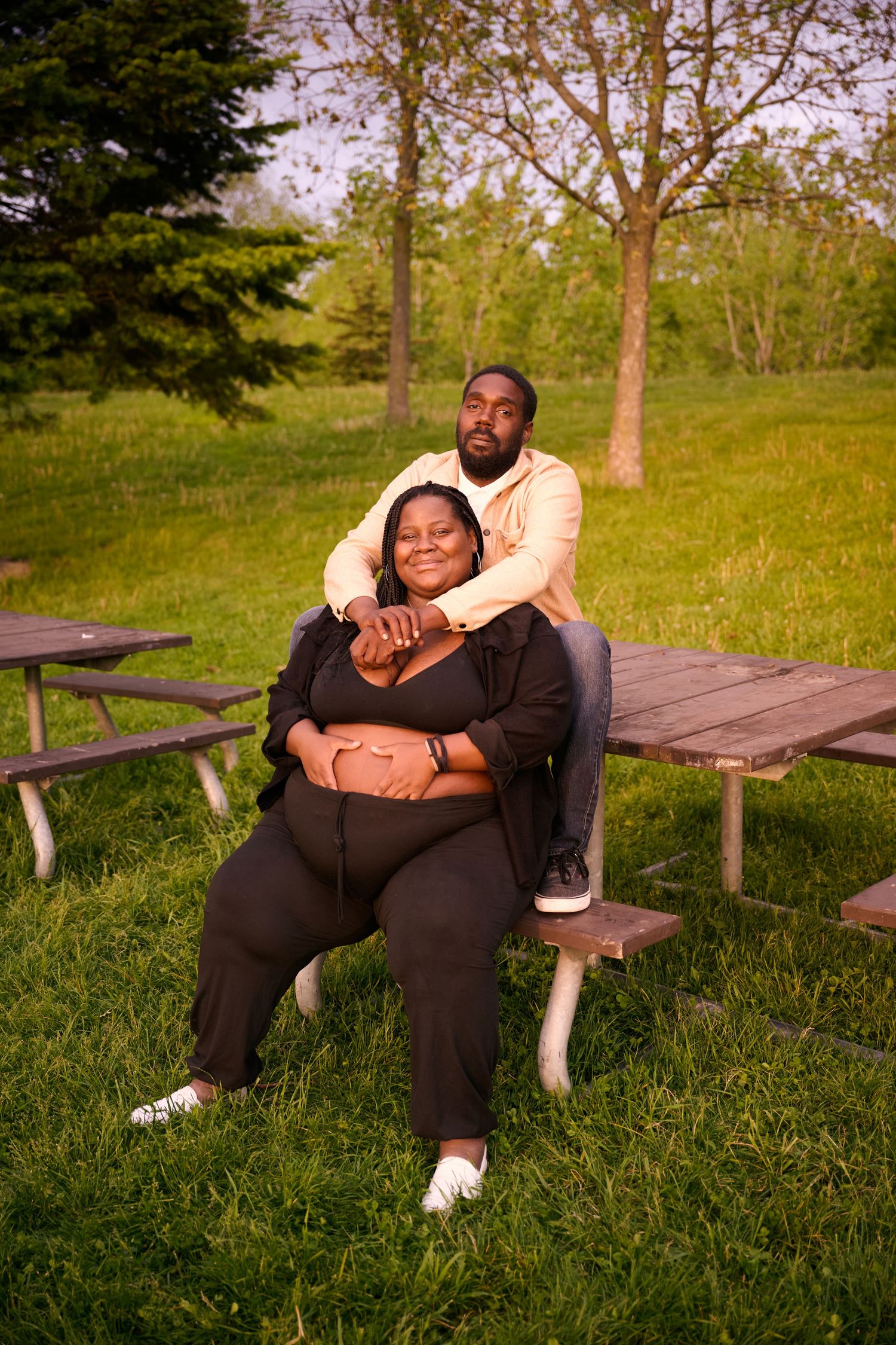 A couple sits together on a wooden picnic bench in a sunny park with green grass and trees in the background.