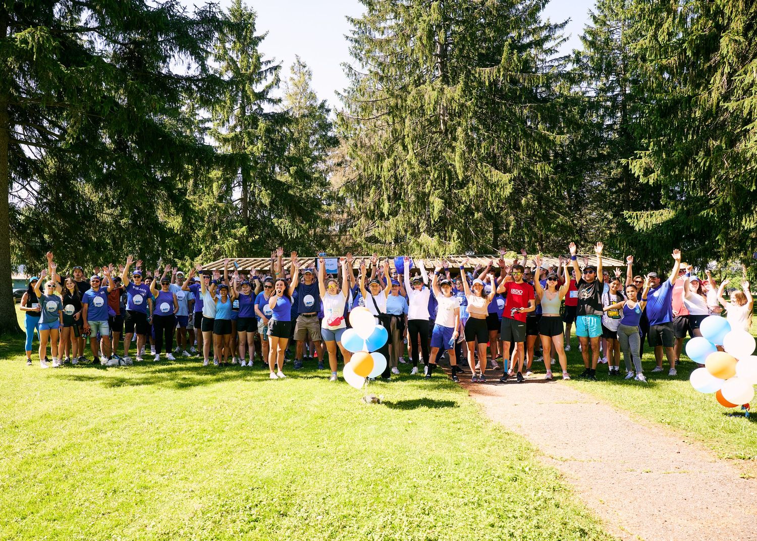 Large group of people celebrating outdoors in a park, raising hands with blue and white balloons on a sunny day.