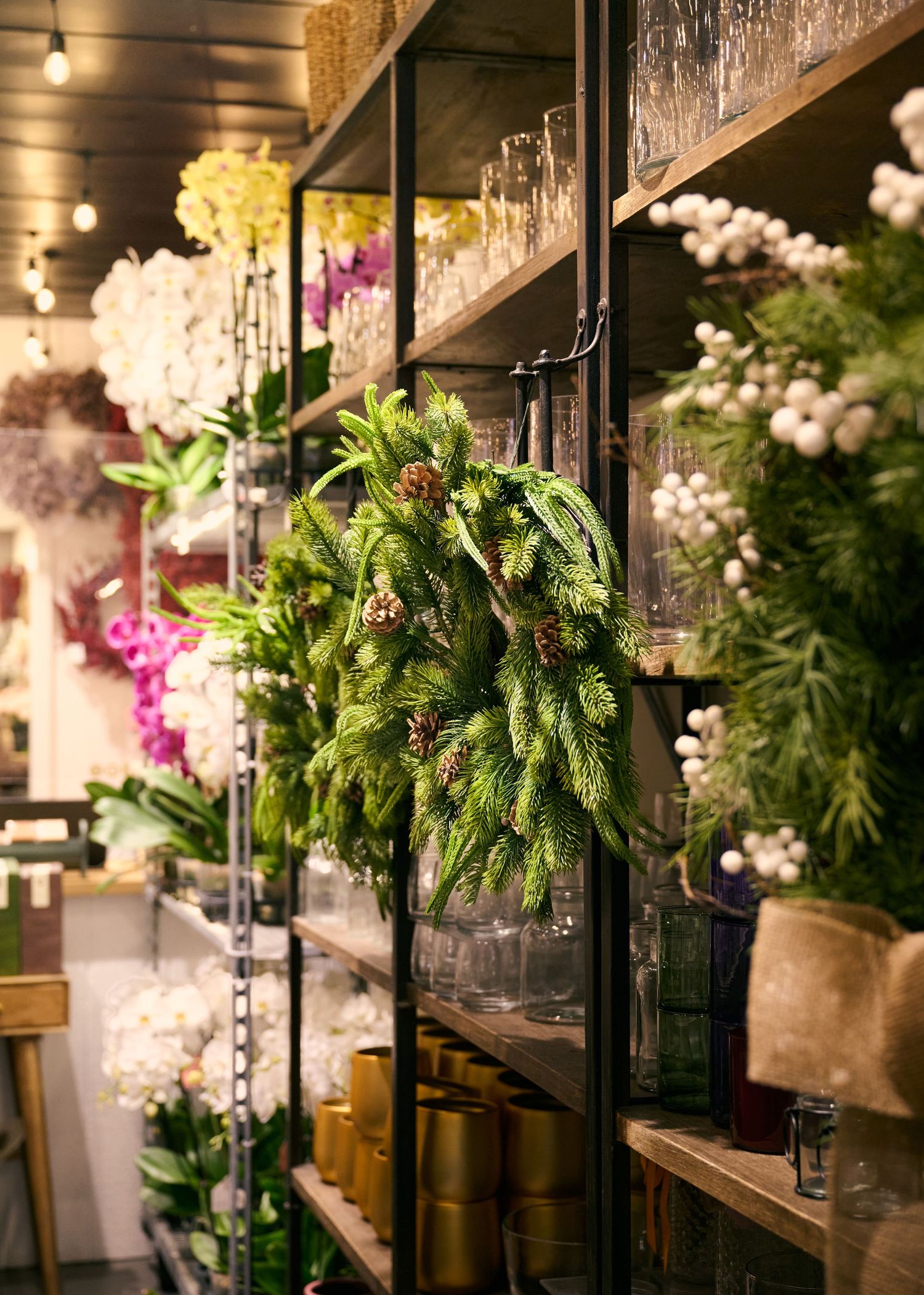 Lush evergreen branches and colorful flowers displayed on dark wooden shelves in a floral shop interior.