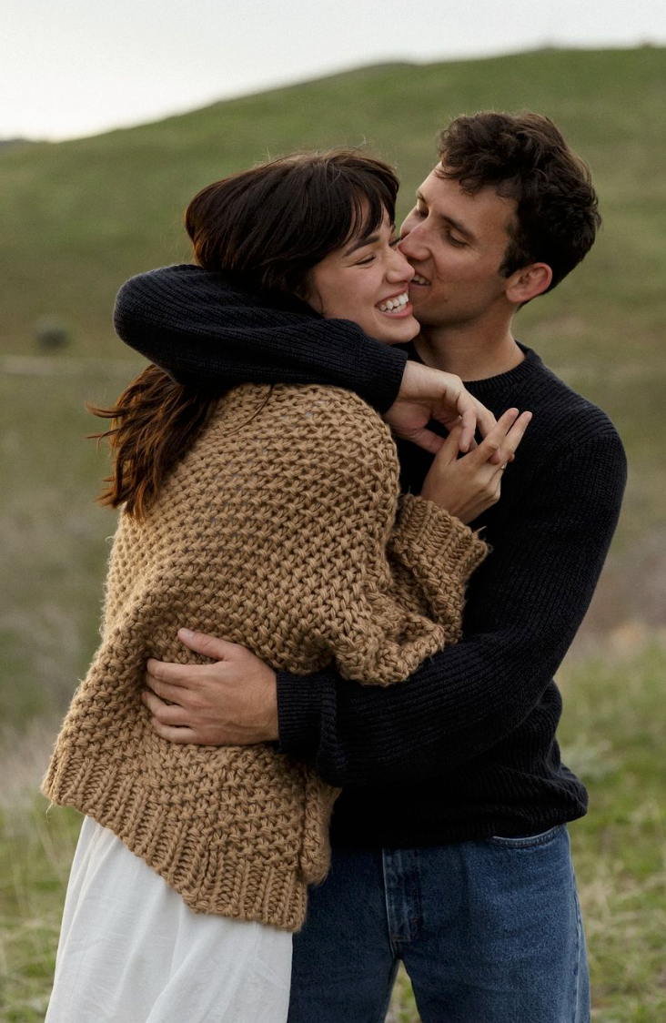 A couple in casual attire shares a romantic moment outdoors with the woman wearing a chunky knit sweater.