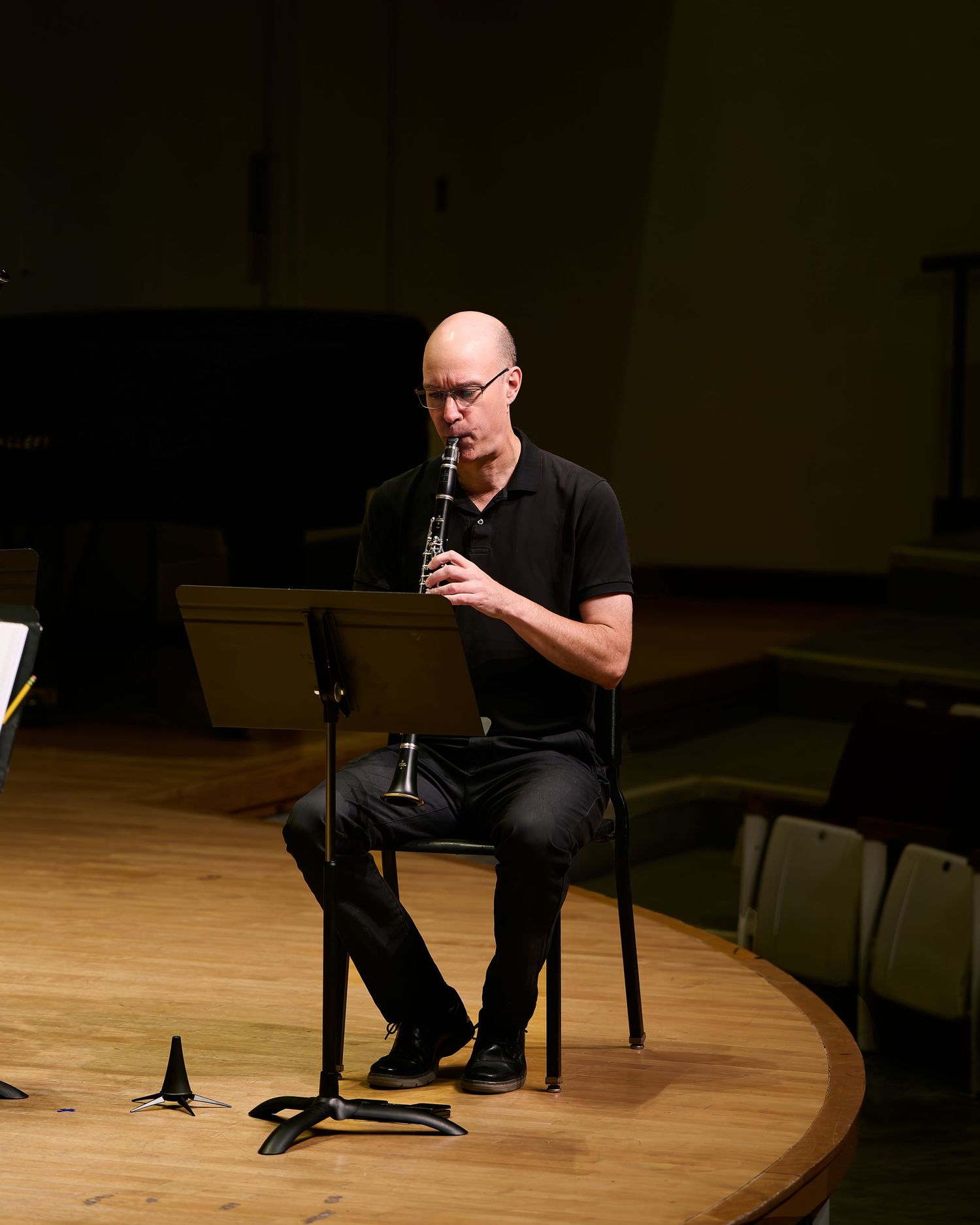 Bald man in black outfit playing clarinet while seated on stage at a music performance.