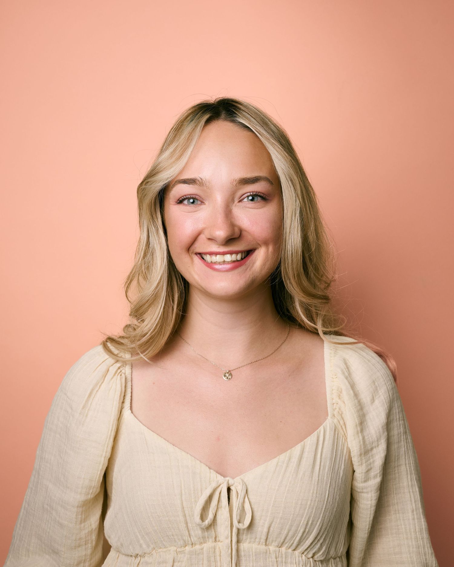Smiling young blonde woman in cream dress poses against a soft peach background for a portrait photo.