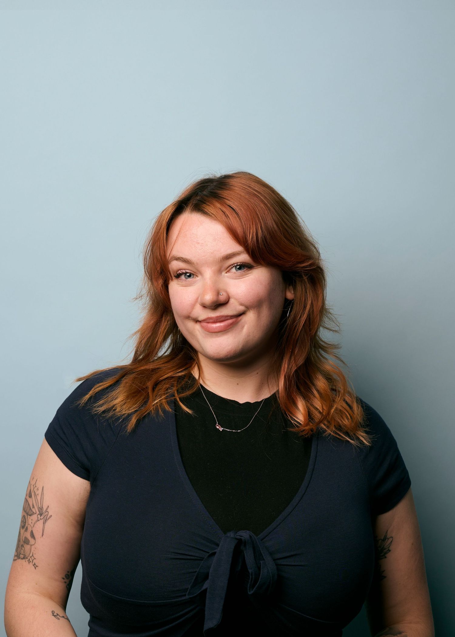 Studio portrait of someone with red wavy hair wearing a black top with tattoo visible on arm against light blue background.