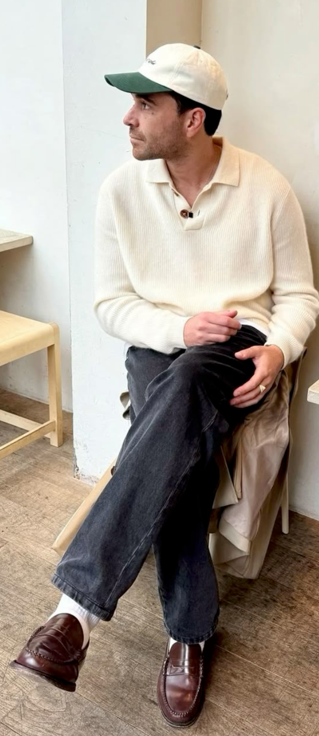 Person wearing cream sweater and dark jeans sits casually in chair wearing brown loafers and baseball cap.
