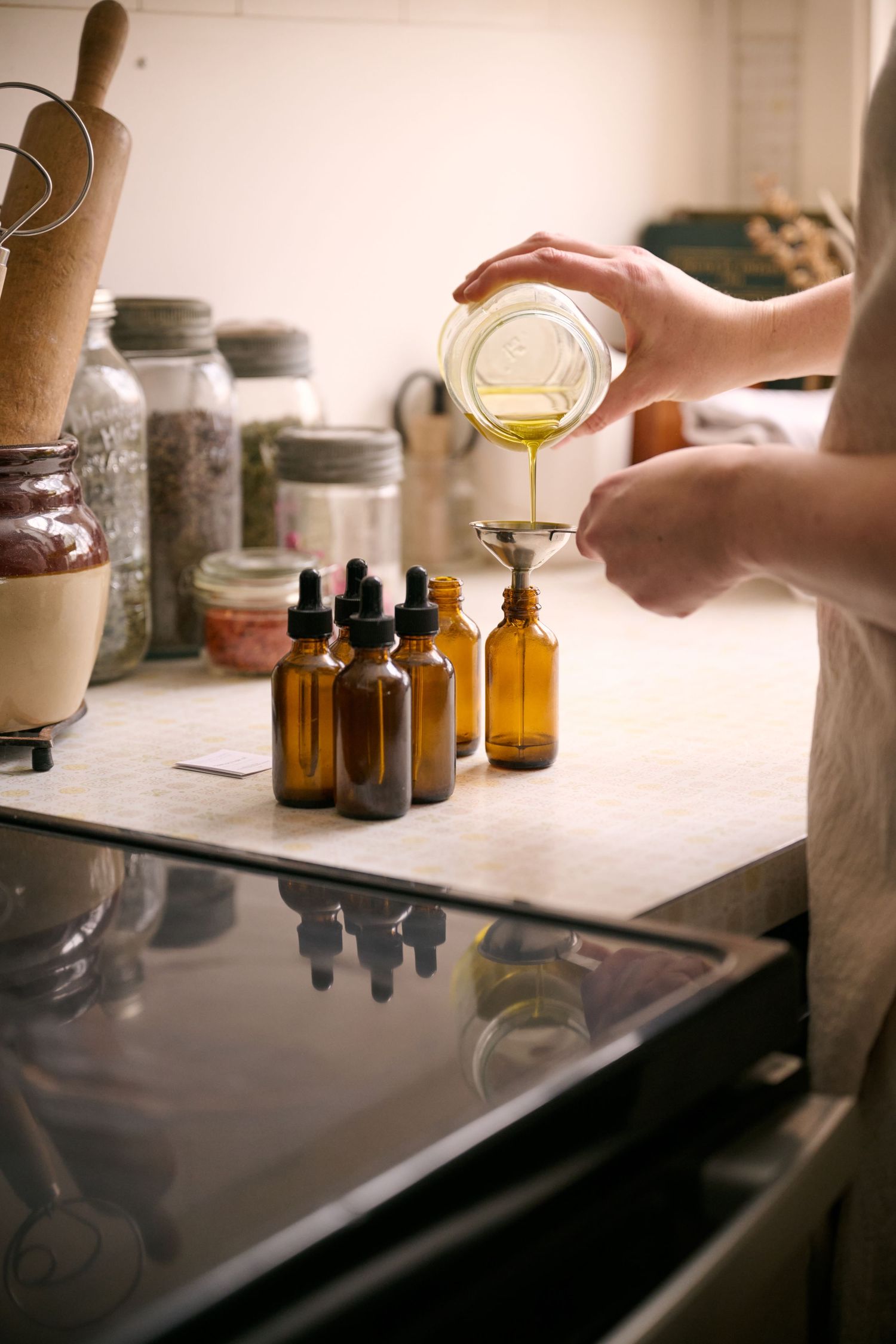 Hands pouring liquid into amber glass bottles on a counter with herbal ingredients and supplies.