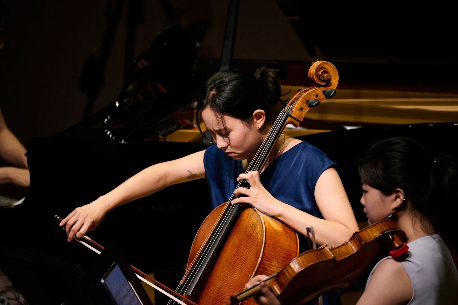 Young female cellist performs passionately on stage during an orchestra concert, with another musician visible behind her.
