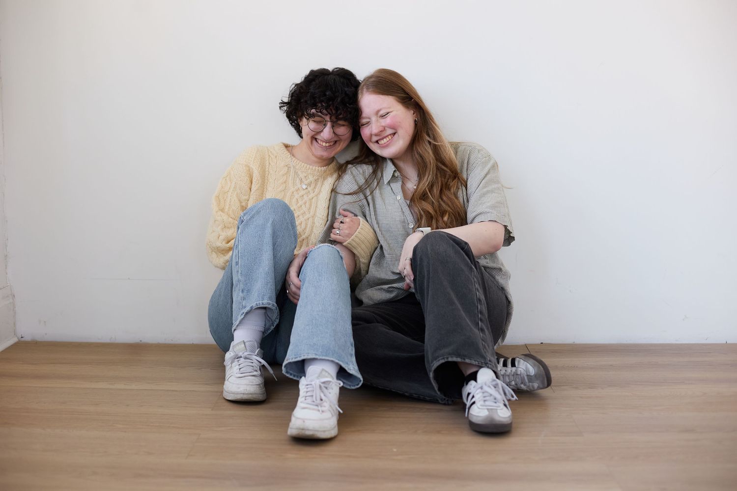 Two people sitting on wooden floor wearing jeans and sneakers lean against white wall while sharing a casual moment.