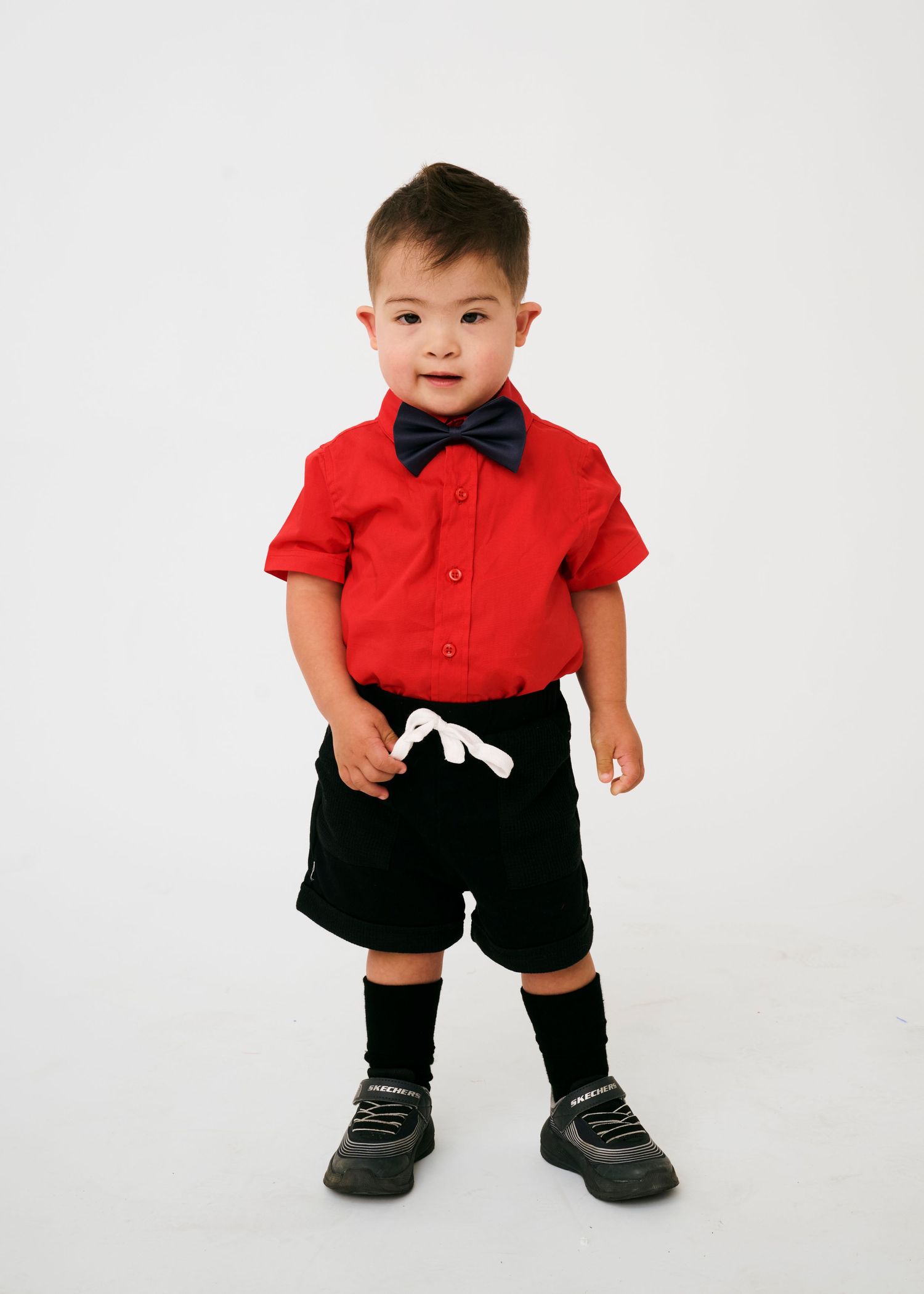 A toddler in a festive red polo shirt and black shorts poses against a white backdrop.