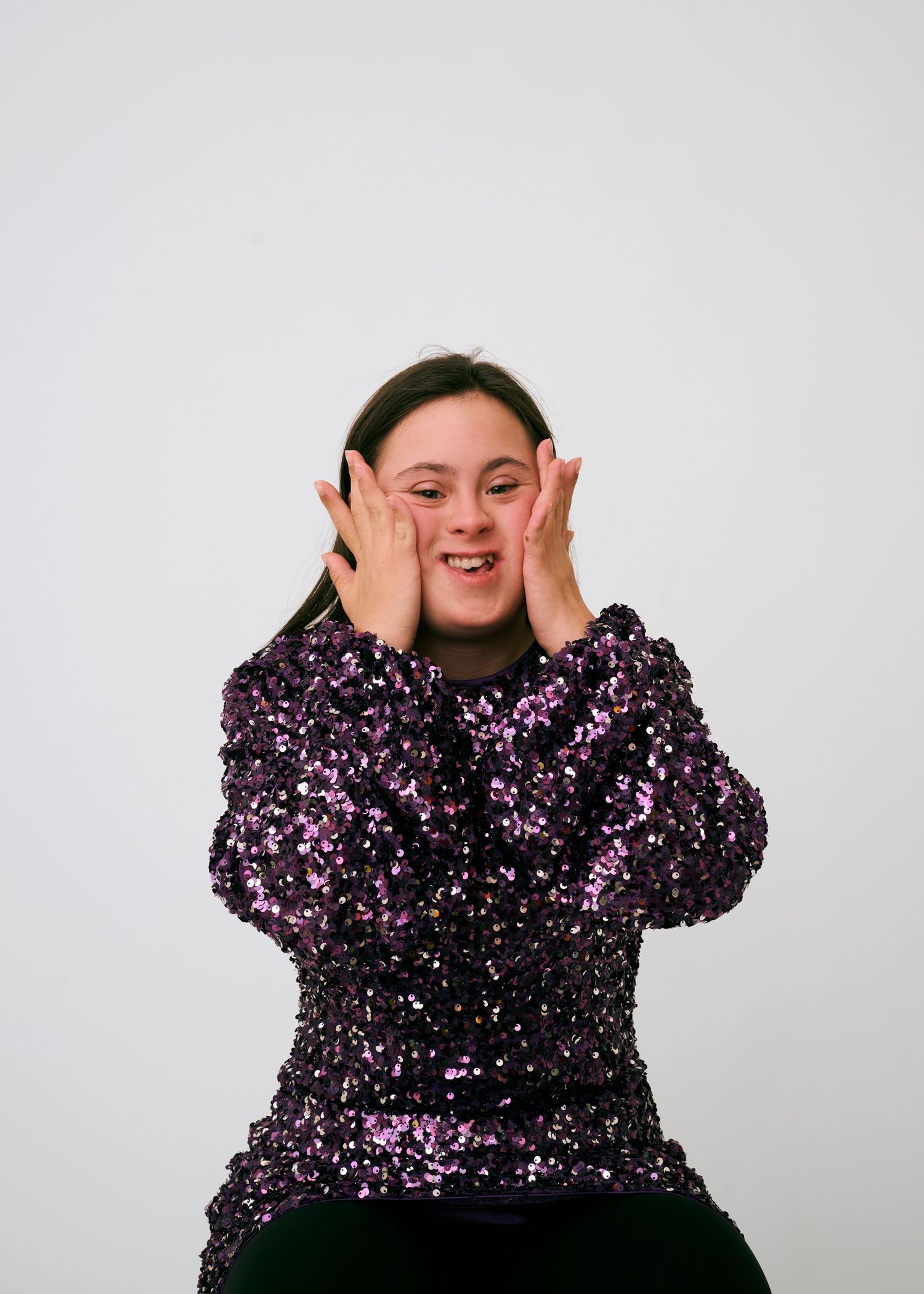Person wearing sparkly purple sequin top poses playfully against white backdrop.