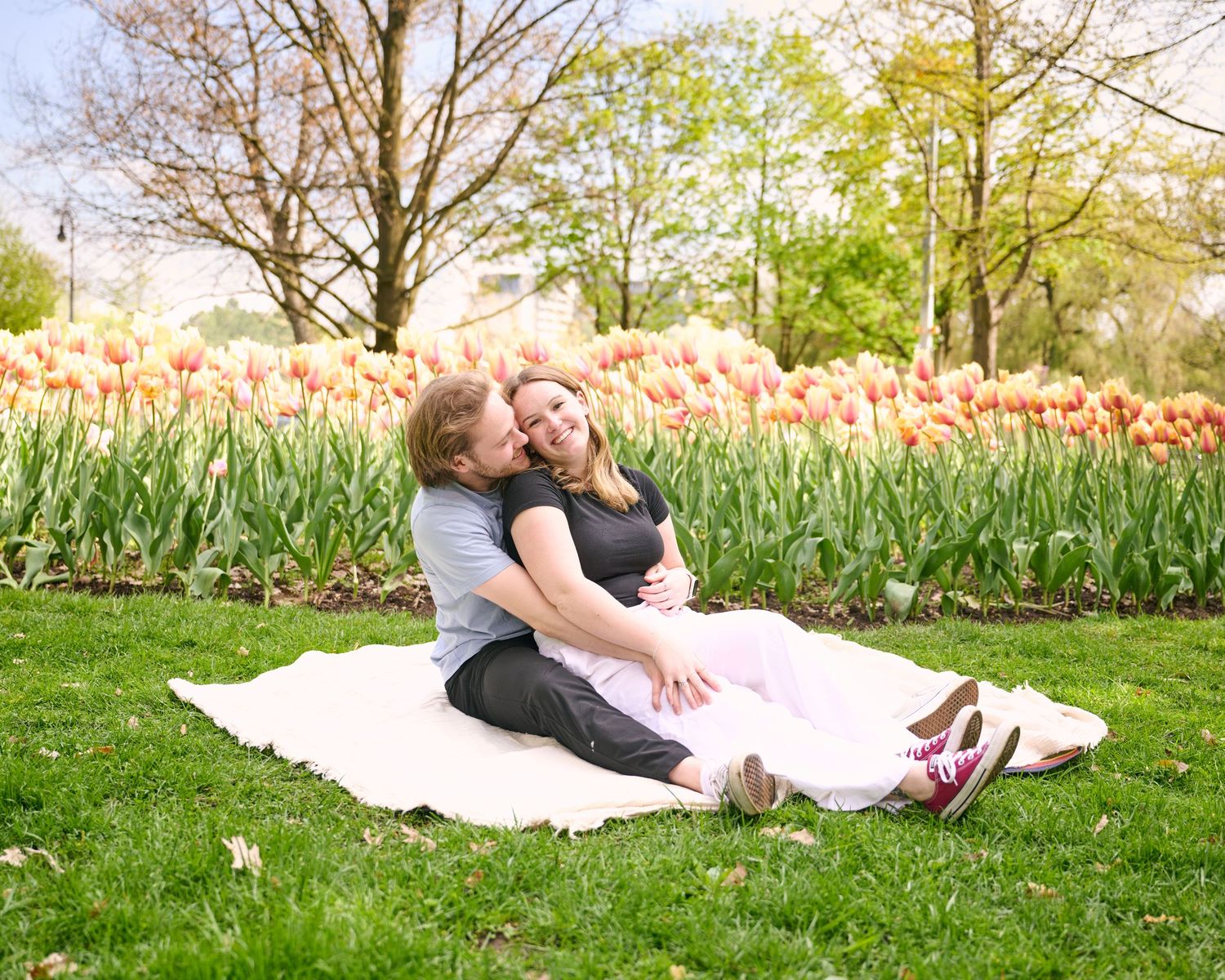 Couple sitting on blanket in park surrounded by blooming pink tulips during a maternity photo session.