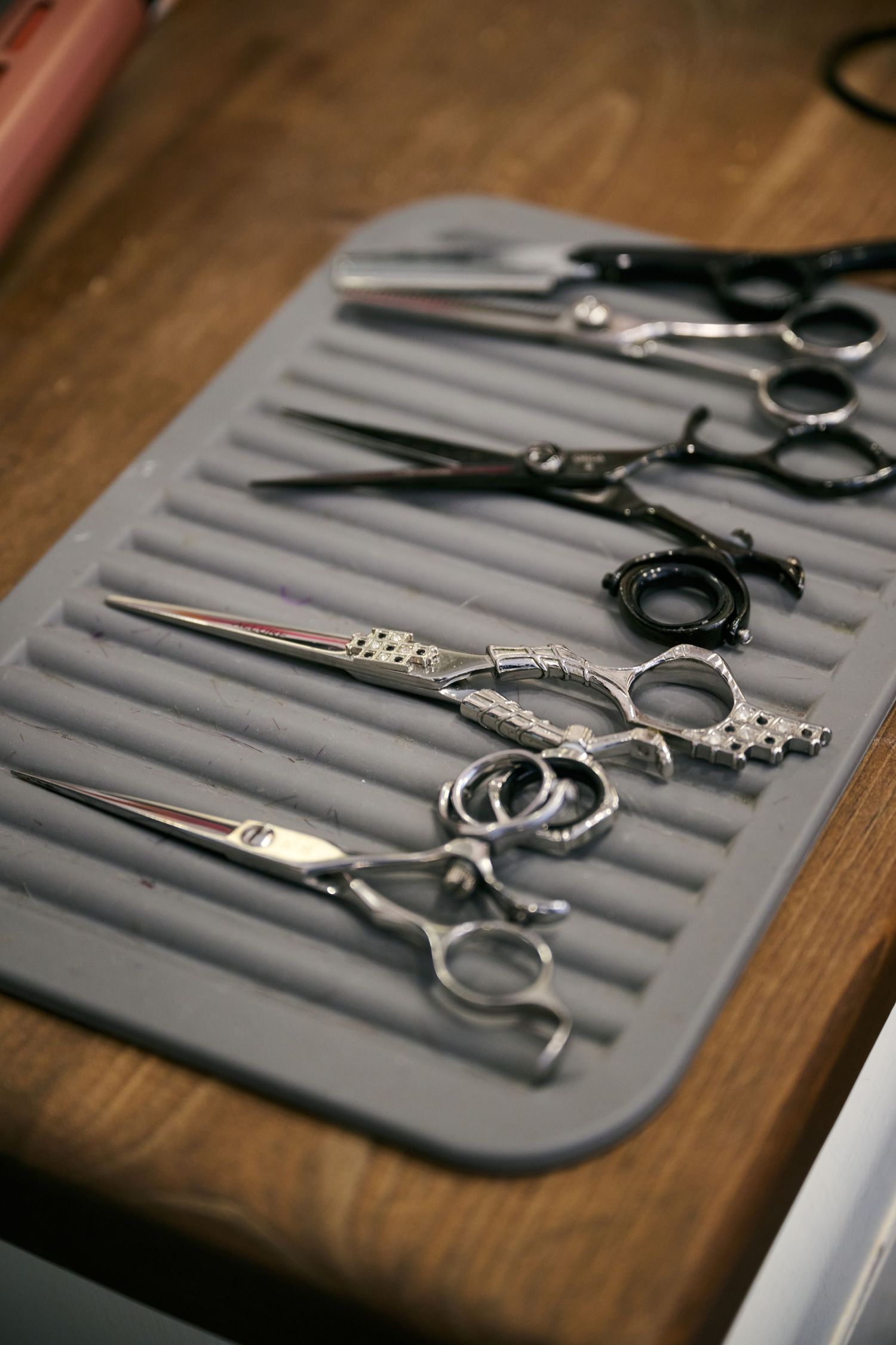 Professional hair cutting scissors and shears arranged on a gray silicone mat on wooden surface in a salon.